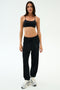 A woman stands against a plain background in SPLITS59’s Andie Oversized Fleece Varsity 59 Sweatpant in Black, paired with a black sports bra and white sneakers—perfect fleece joggers for cool weather workouts. Made in Los Angeles.