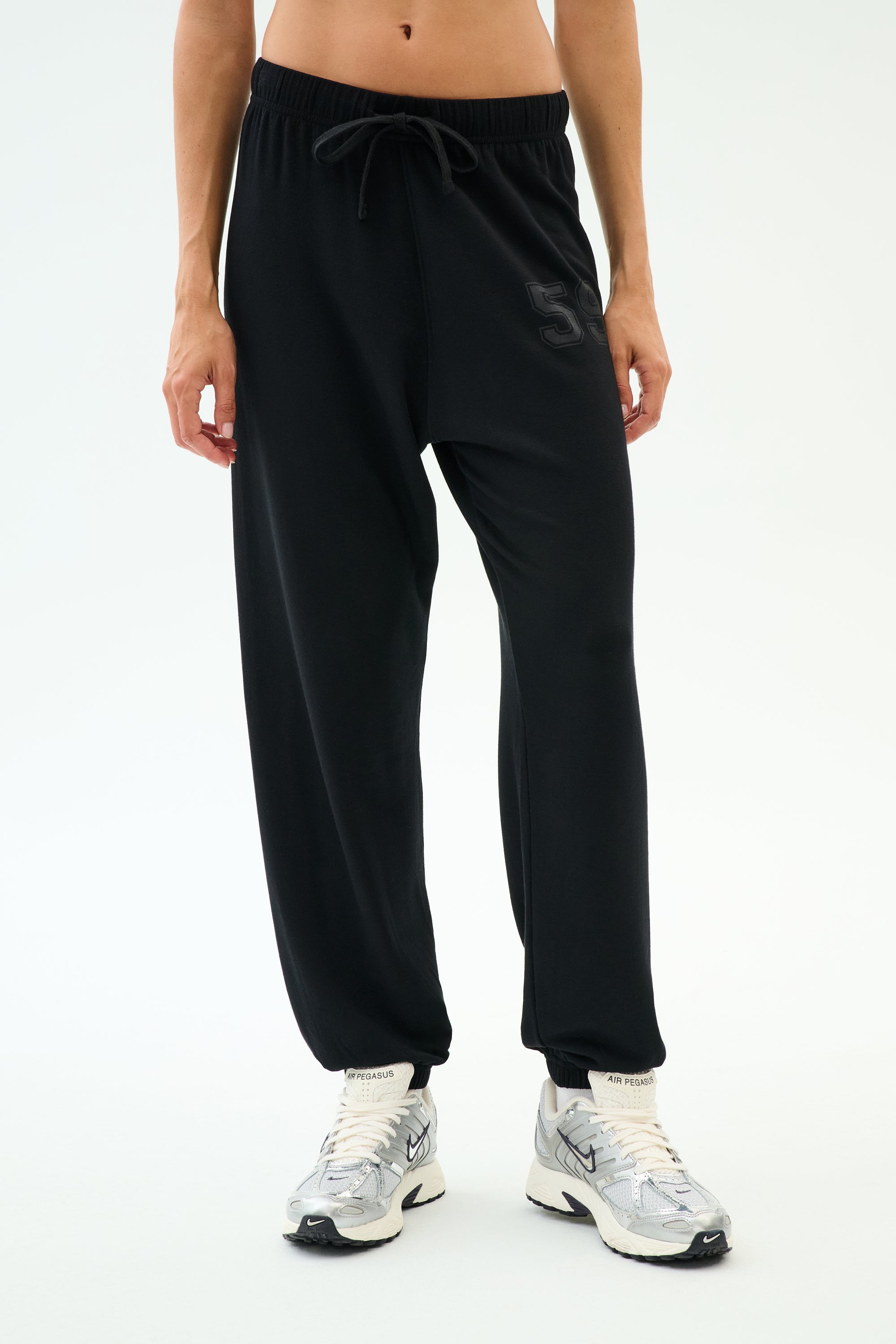 A person wears SPLITS59's Andie Oversized Fleece Varsity 59 Sweatpant in black, featuring a "59" on the left thigh, paired with white sneakers—ideal for cool weather workouts—standing against a plain white background.