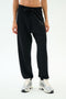 A person wears SPLITS59's Andie Oversized Fleece Varsity 59 Sweatpant in black, featuring a "59" on the left thigh, paired with white sneakers—ideal for cool weather workouts—standing against a plain white background.