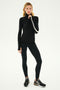 A person stands against a plain background wearing a black and white Jackson Rib Full Length Turtleneck by SPLITS59 with white stripes on the sleeves, black leggings, and black New Balance sneakers, ready for yoga or running.