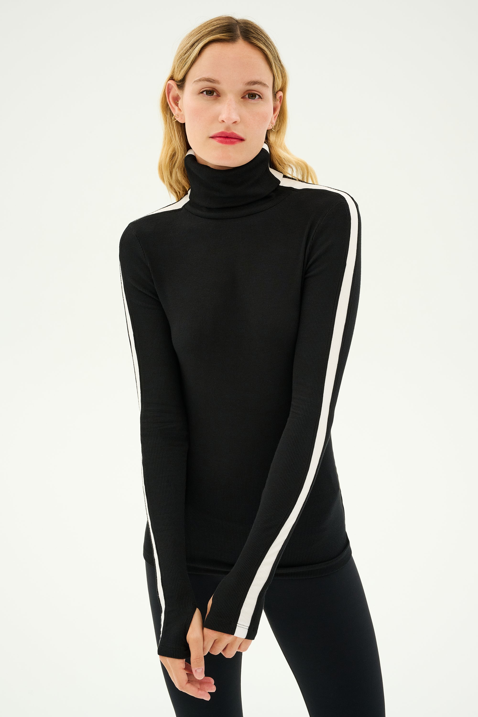 A person with long hair is standing against a plain background, wearing the Jackson Rib Full Length Turtleneck - Black/White by SPLITS59, detailed with white stripes down the sleeves and exuding a serene yoga vibe.