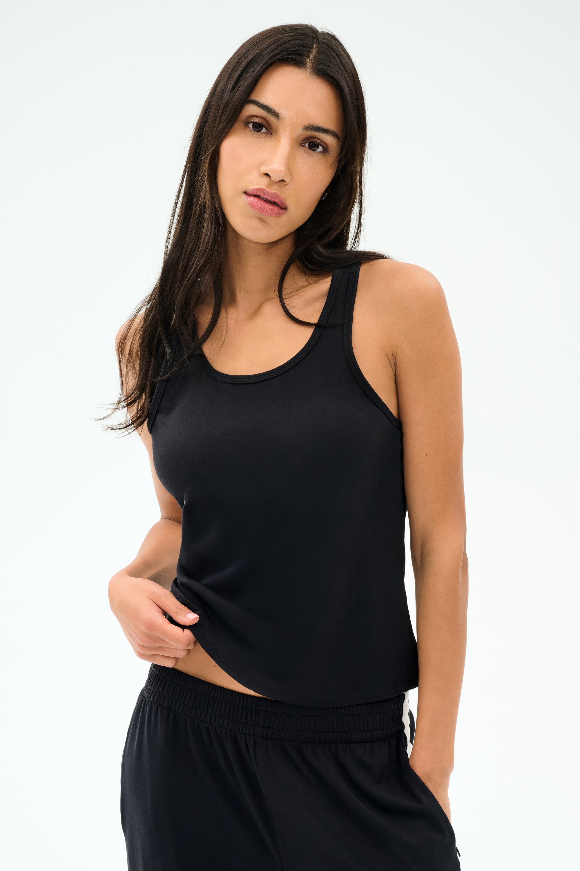 Woman with long dark hair wears the SPLITS59 Mel Airweight Lite Tank in black, paired with black pants, standing against a light background.