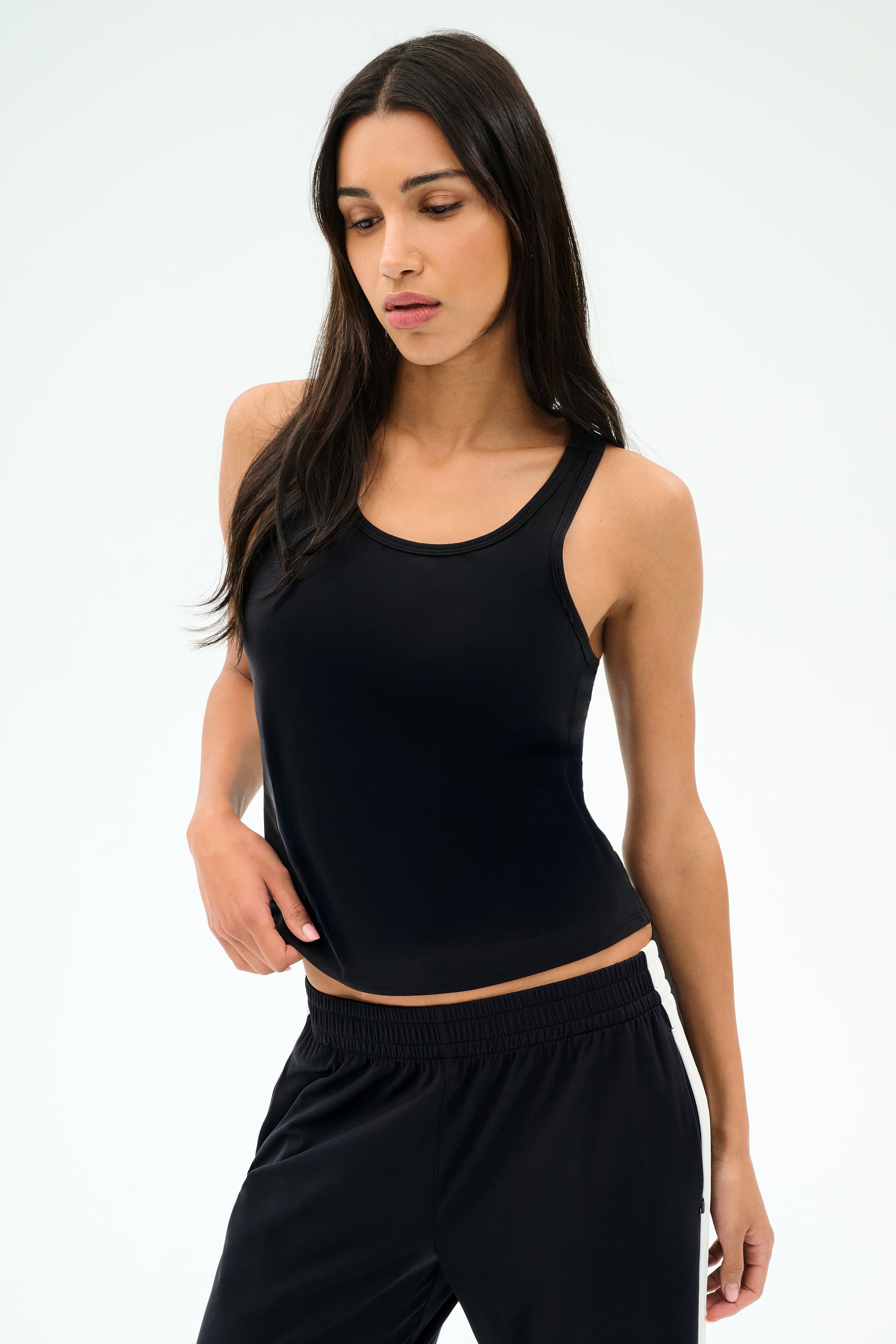 A woman with long dark hair wears the SPLITS59 Mel Airweight Lite Tank in black and black pants, standing against a plain light background.