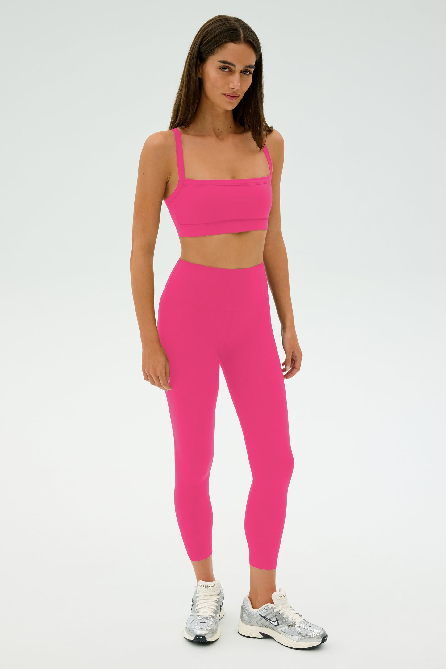 A woman stands facing forward in a matching SPLITS59 Monah Rigor Bra - Berry and high-waisted leggings, ideal for gym workouts, styled with white sneakers against a plain white background.