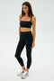 A person in a SPLITS59 Monah Rigor Bra - Black and black leggings stands against a plain background, with long hair and wearing white and gray sneakers, ready for gym workouts.