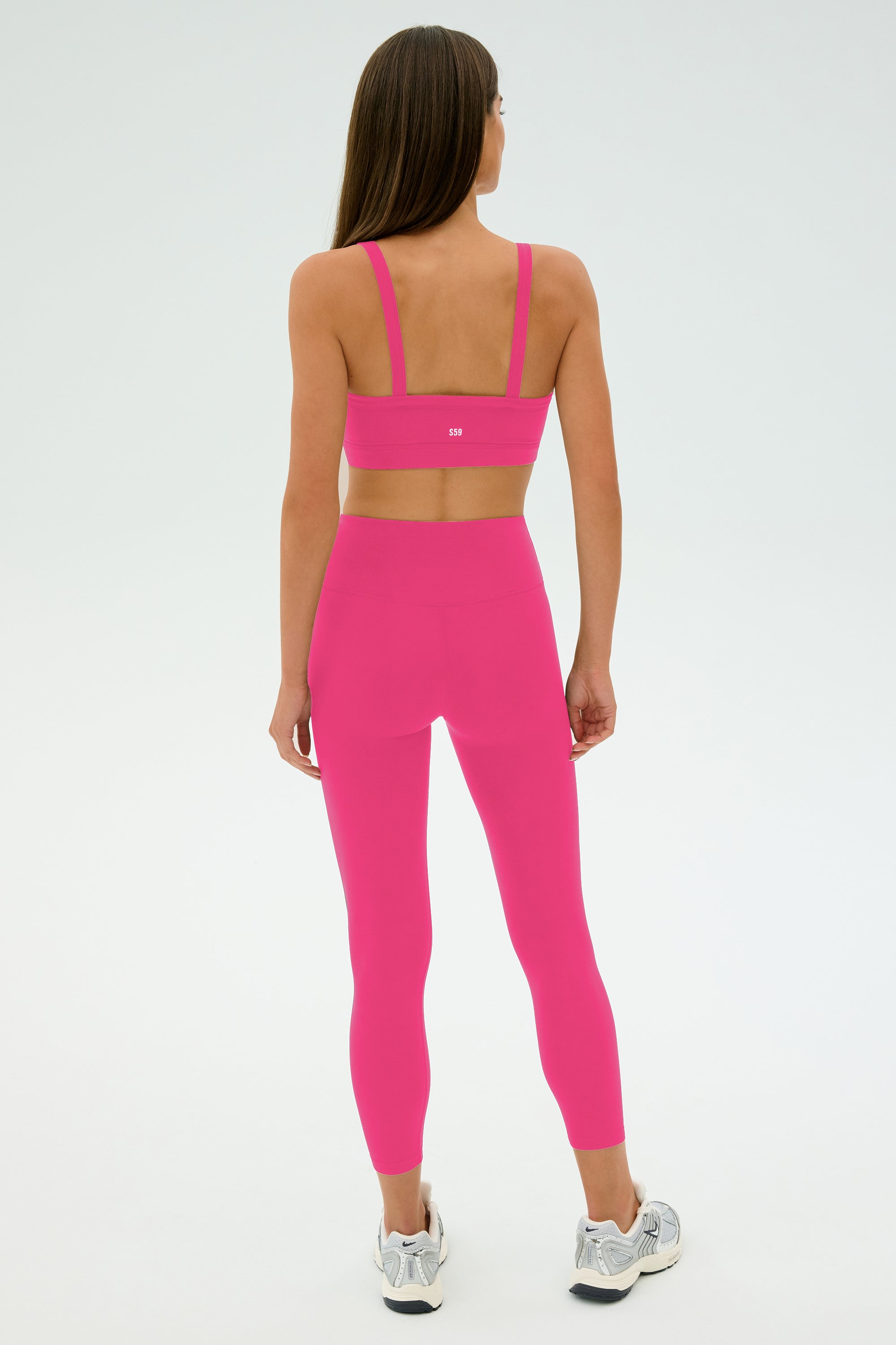 A woman faces away, wearing the SPLITS59 Monah Rigor Bra - Berry with matching pink leggings and white sneakers—ideal for gym workouts—set against a plain light background.