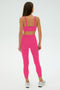 A woman faces away, wearing the SPLITS59 Monah Rigor Bra - Berry with matching pink leggings and white sneakers—ideal for gym workouts—set against a plain light background.