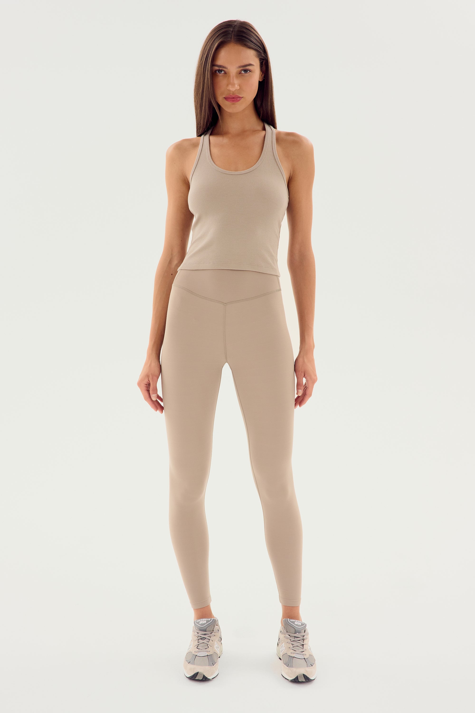 A woman stands against a white background wearing a beige tank top, high waist Airweight Super High Waist 7/8 - Latte leggings made from ultra luxe Airweight fabric, and gray sneakers by SPLITS59.