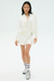 A woman stands against a white background wearing the SPLITS59 Airweight Bomber Jacket in white, a matching dress, striped socks, and silver sneakers.