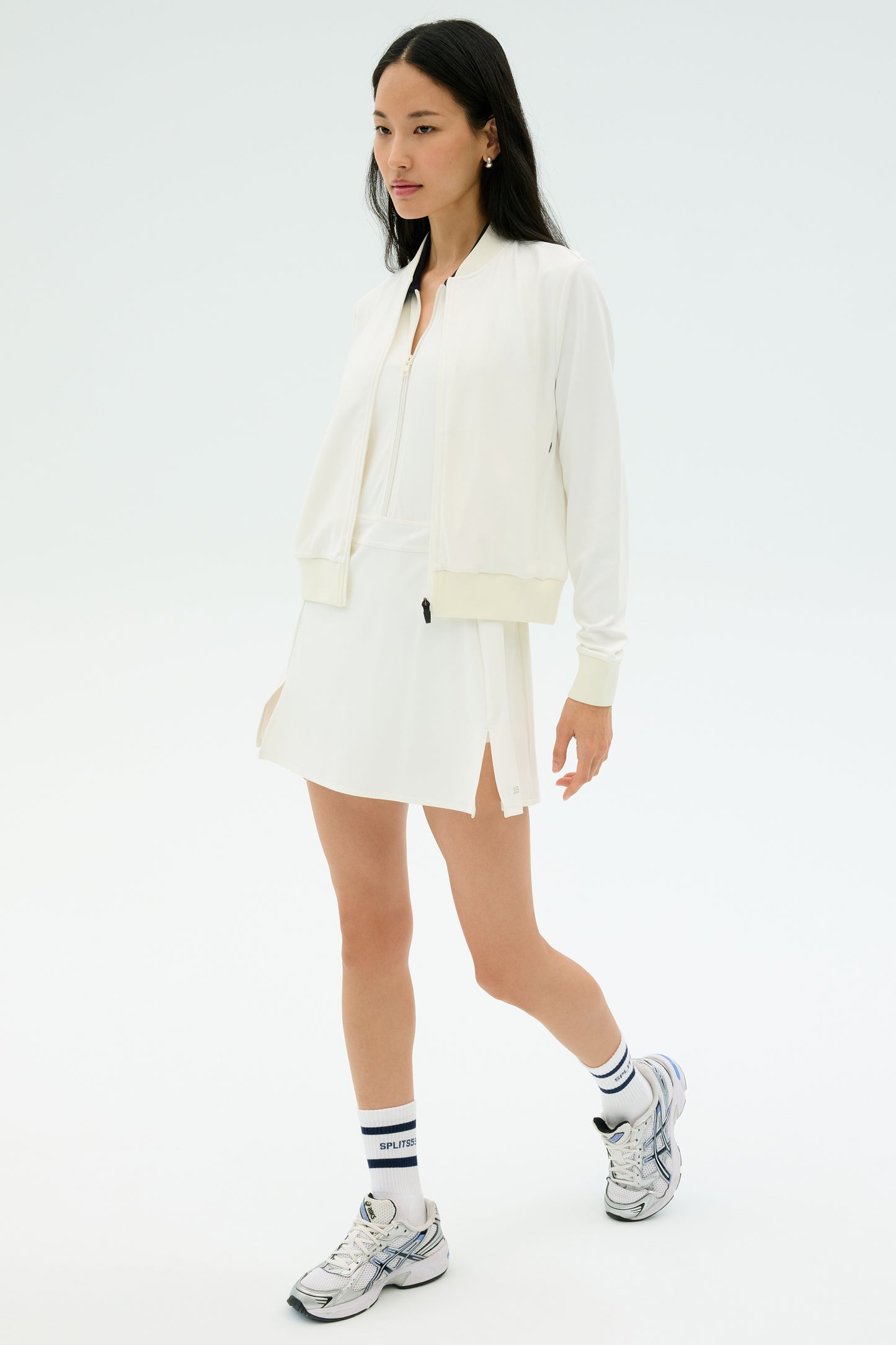 A woman wears the SPLITS59 Airweight Bomber Jacket in white, paired with a white dress, white socks with blue stripes, and silver athletic shoes against a plain light background.