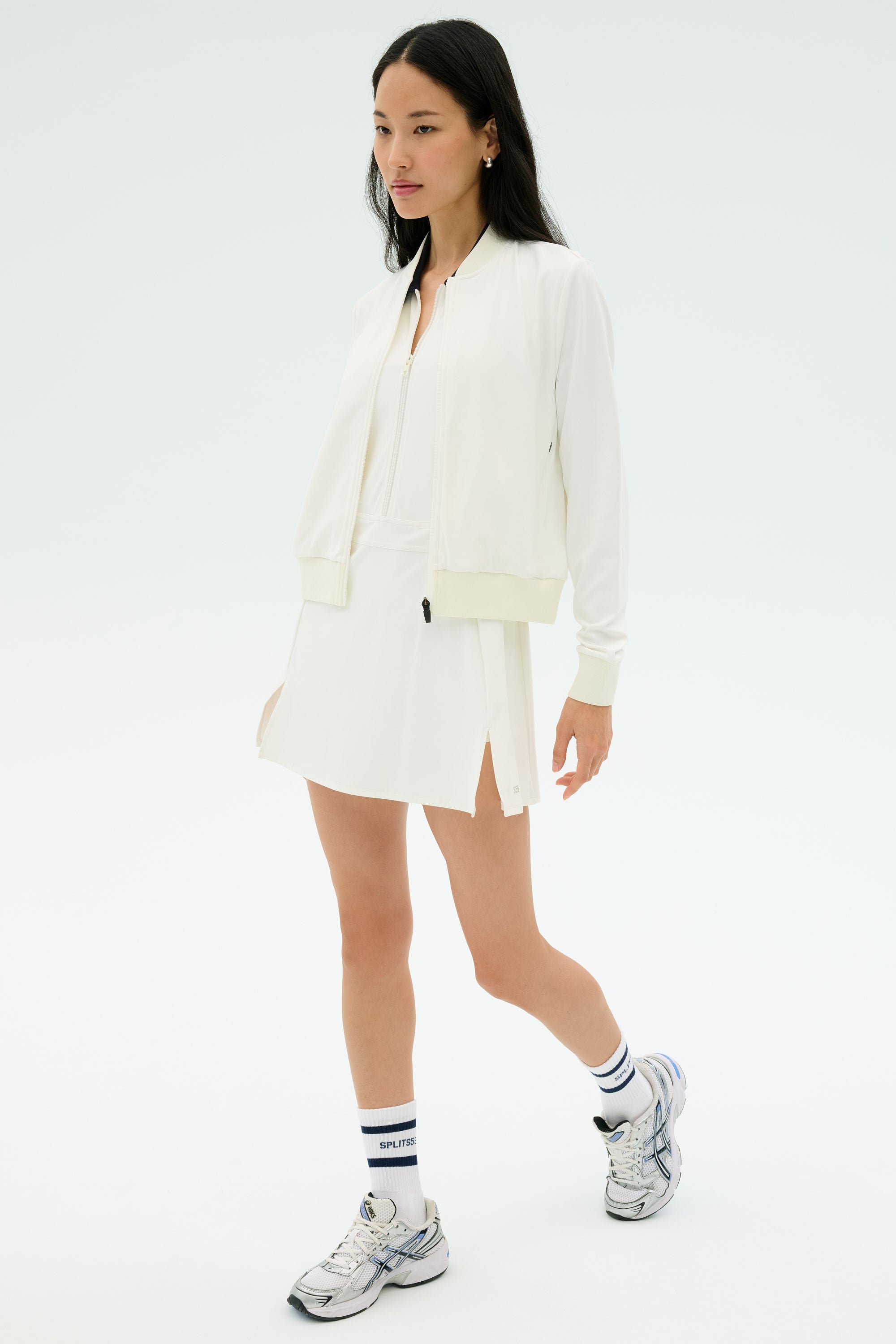 A woman wears the SPLITS59 Airweight Bomber Jacket in white, paired with a white dress, white socks with blue stripes, and silver athletic shoes against a plain light background.