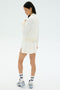 A woman faces slightly away against a plain white background, wearing the SPLITS59 Airweight Bomber Jacket in white, paired with a white skirt, striped white socks, and silver sneakers.