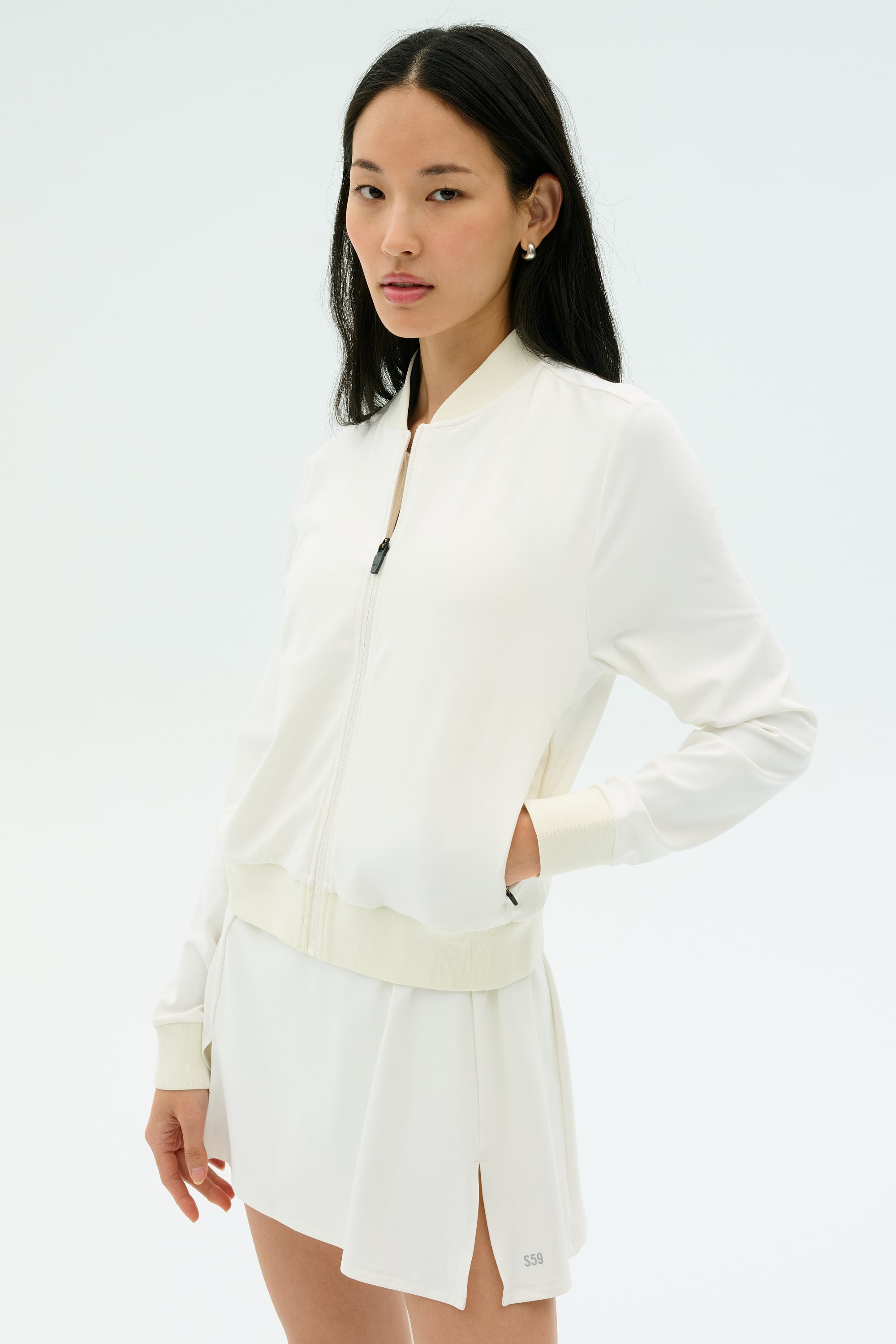 A woman stands against a plain background wearing the SPLITS59 Airweight Bomber Jacket in white with her hands in the jacket pockets, paired with a matching white skirt.