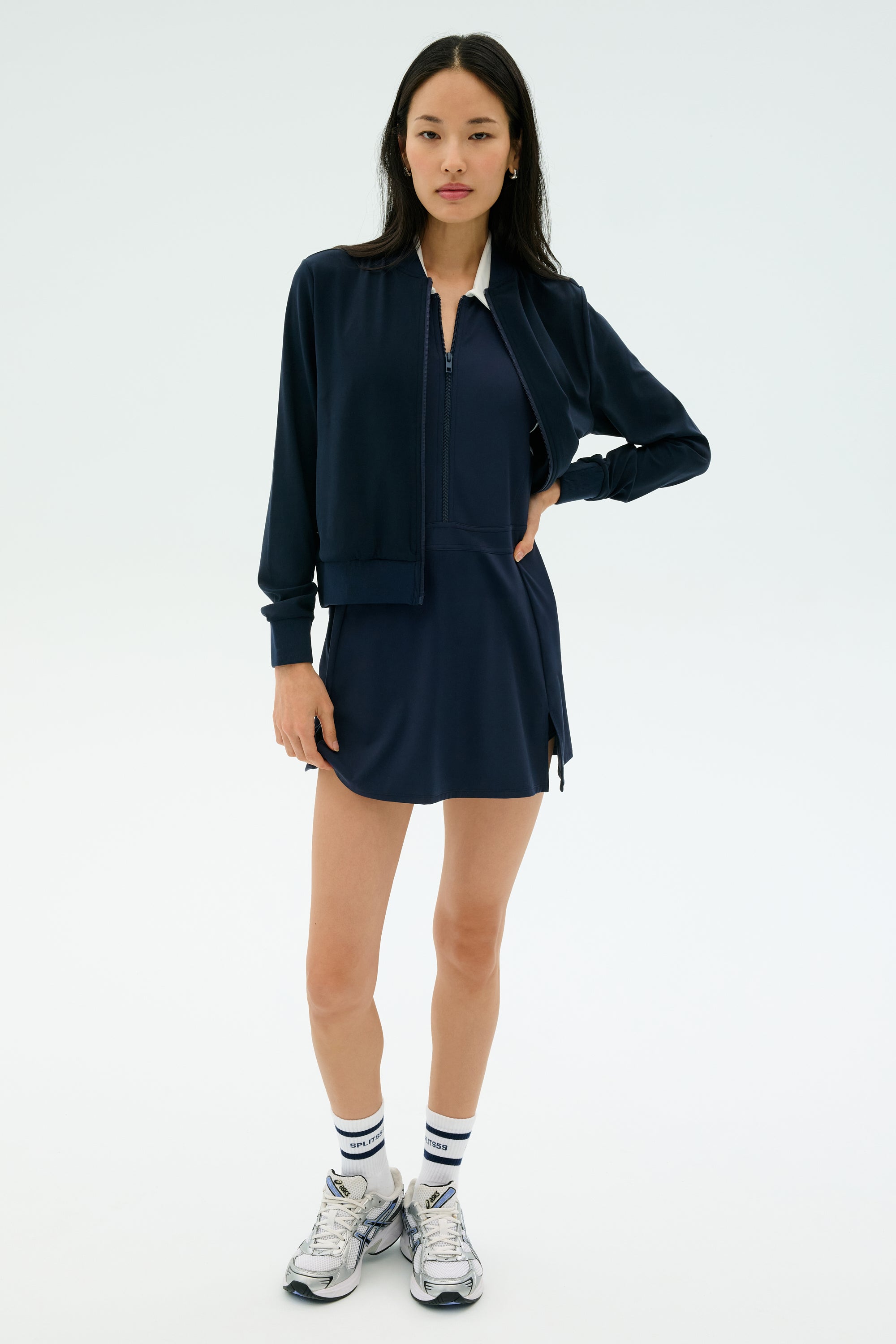 A woman faces forward wearing the SPLITS59 Airweight Bomber Jacket in Indigo, paired with a matching dress, white socks with blue stripes, and silver athletic sneakers against a plain light background.