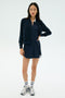 A woman faces forward wearing the SPLITS59 Airweight Bomber Jacket in Indigo, paired with a matching dress, white socks with blue stripes, and silver athletic sneakers against a plain light background.