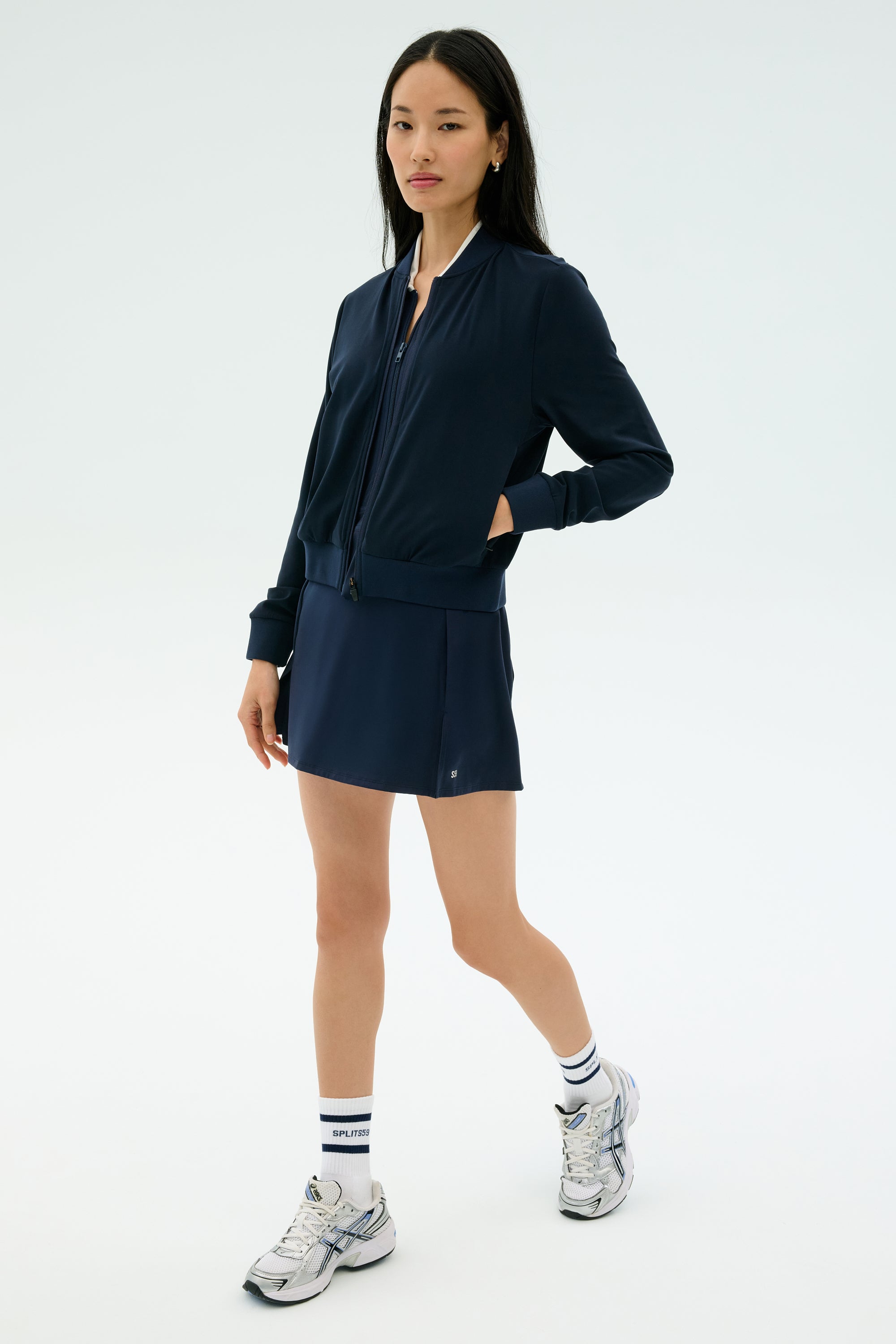 A woman stands against a plain light background wearing the SPLITS59 Airweight Bomber Jacket in Indigo, paired with a matching navy skirt, white socks, and silver athletic sneakers.
