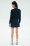 A woman stands facing away, wearing the SPLITS59 Airweight Bomber Jacket in Indigo, a navy pleated skirt, striped white socks, and white athletic shoes against a plain light background.