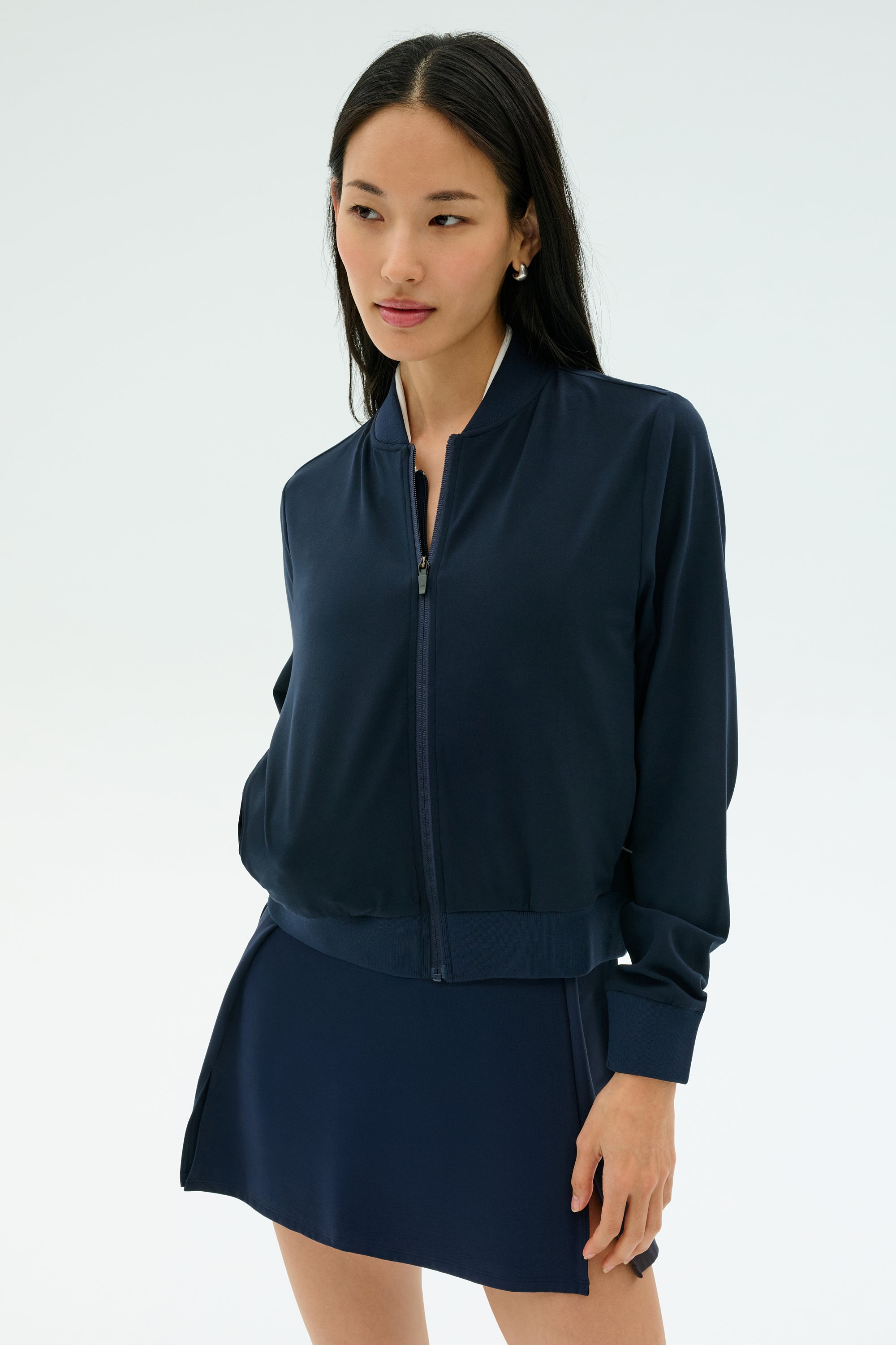 A woman stands against a plain background wearing the SPLITS59 Airweight Bomber Jacket in Indigo, paired with a matching skirt, with her hands relaxed at her sides.