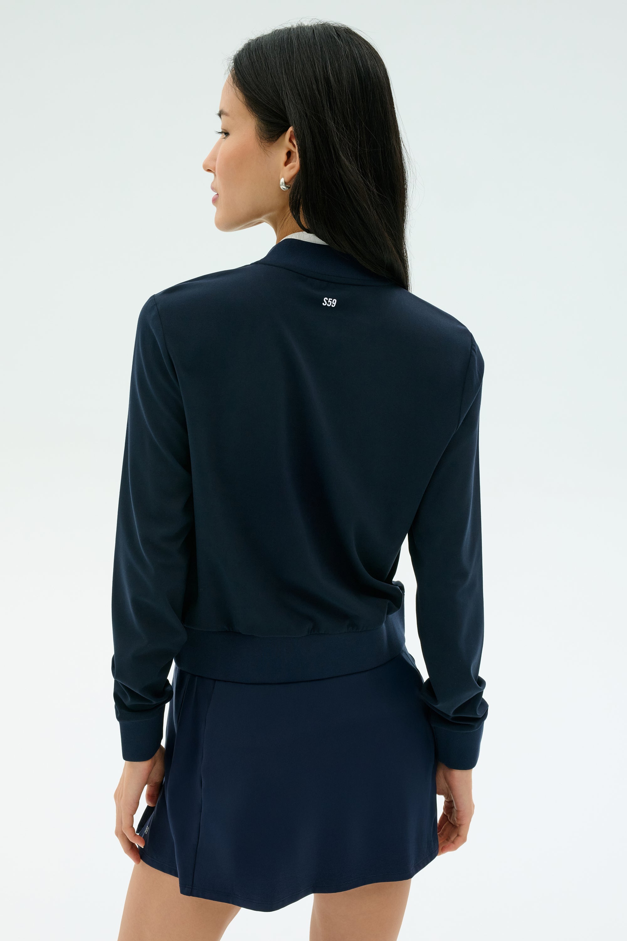 A woman faces away from the camera, wearing the SPLITS59 Airweight Bomber Jacket in Indigo, paired with a matching skirt against a plain light background.