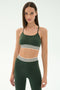 A woman wearing the SPLITS59 Cali Active Rib Bra in Heather Military with matching high-waisted leggings featuring white and black striped bands stands against a plain light background.