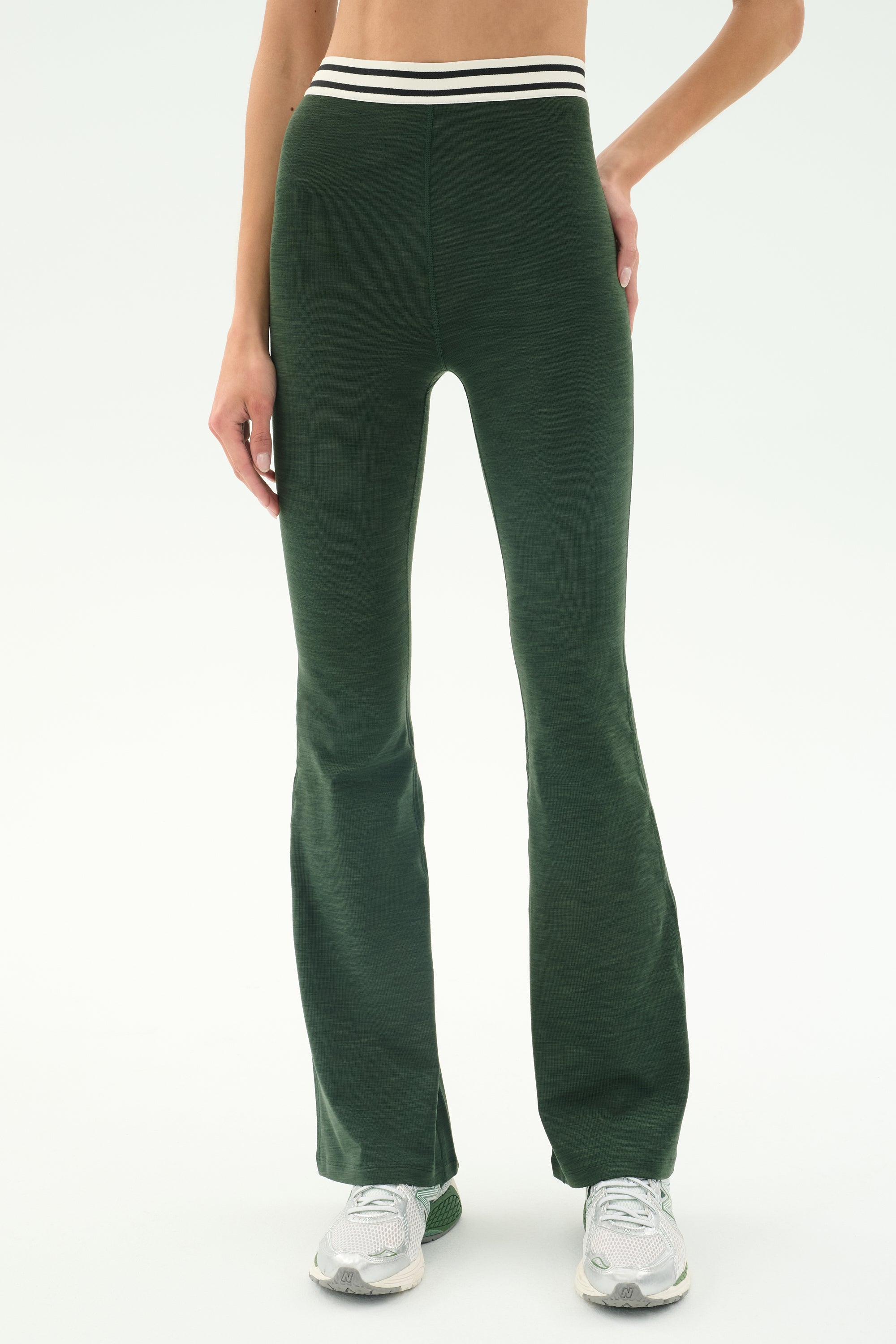 A person wears SPLITS59's Raquel Active Rib Flare - Heather Military pants with a white and black striped waistband and white sneakers, standing against a plain light background.