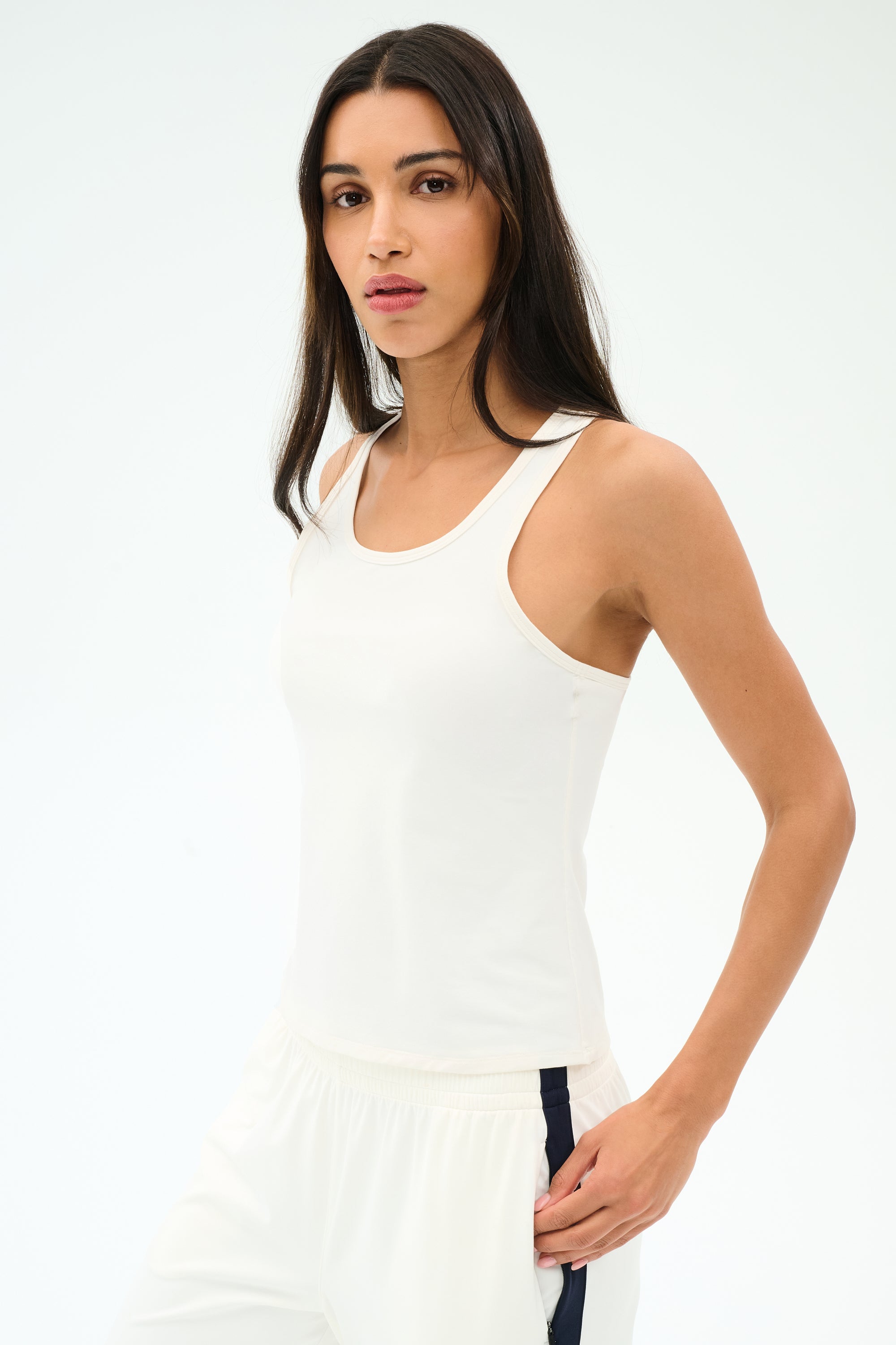 A woman with long dark hair wears the SPLITS59 Mel Airweight Lite Tank in white, paired with white pants featuring a navy stripe, standing against a plain light background.
