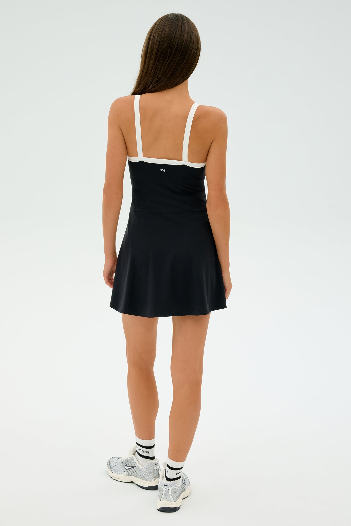 A person wears the SPLITS59 Monah Rigor Dress - Black/White featuring stretchy spandex and white straps, paired with white socks and silver sneakers, standing away in a bright studio setting.