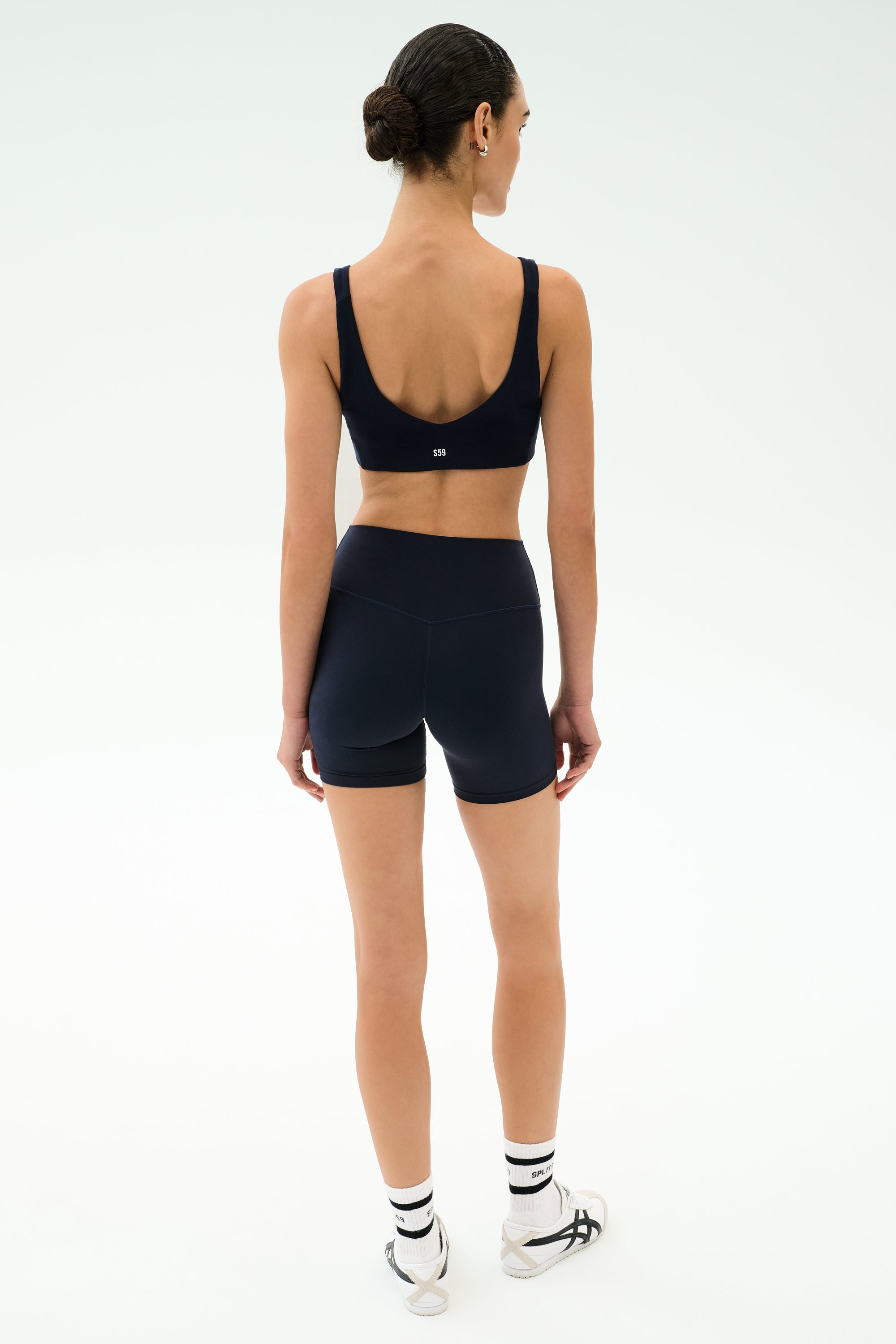 A woman faces away, wearing SPLITS59 Airweight High Waist Short in Indigo with a navy seamless workout top, white socks, and white sneakers against a plain light background.