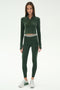A person faces forward wearing the SPLITS59 Stevie Active Rib Half Zip in Heather Military, paired with matching high-waisted leggings and white sneakers, against a plain white background.