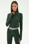 A woman wears the SPLITS59 Stevie Active Rib Half Zip in Heather Military, a long-sleeve zip-up top paired with high-waisted leggings, standing against a plain light background.