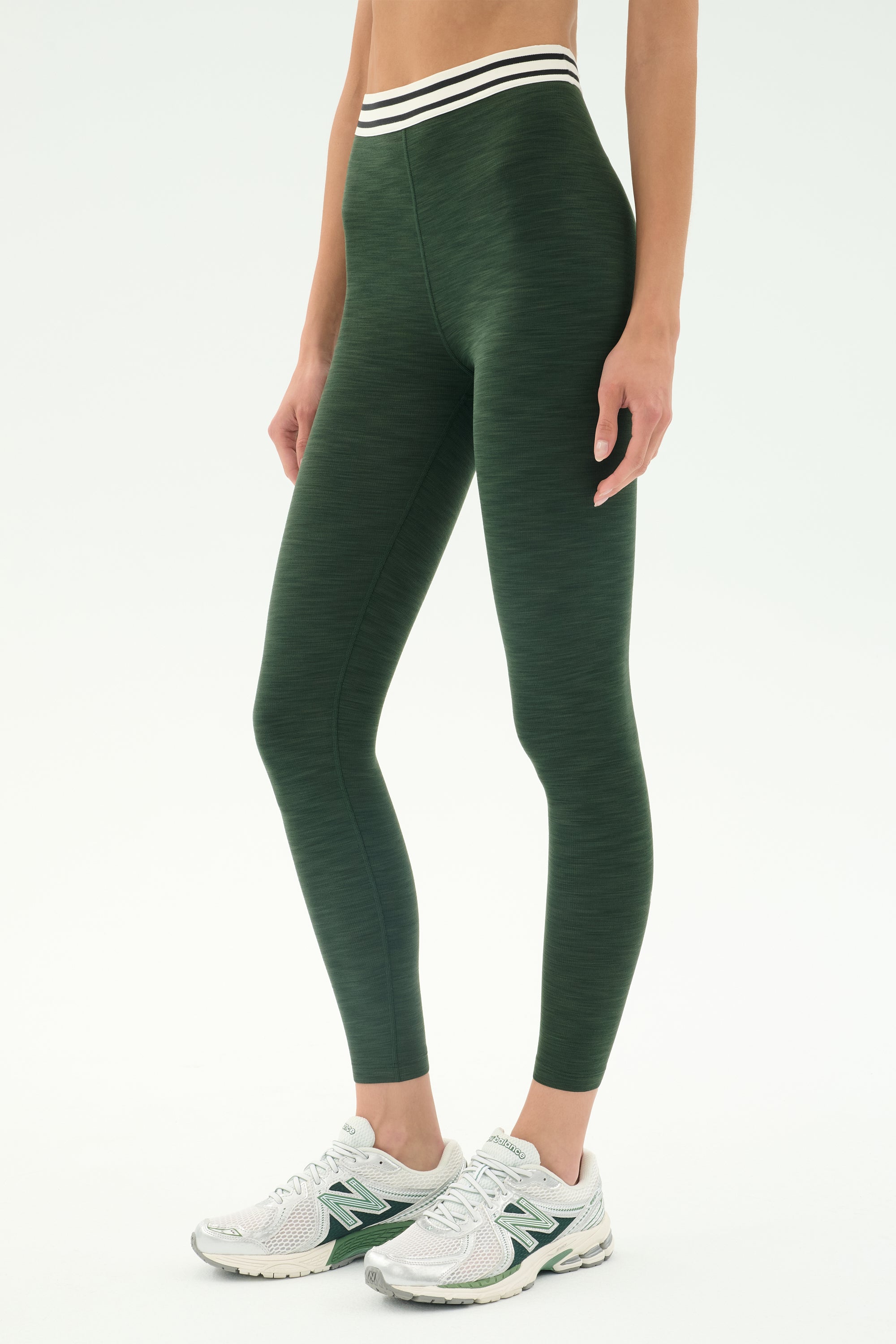 A person wears SPLITS59 Bailey Active Rib High Waist 7/8 leggings in Heather Military, featuring a striped waistband, paired with white athletic shoes, standing against a plain background.