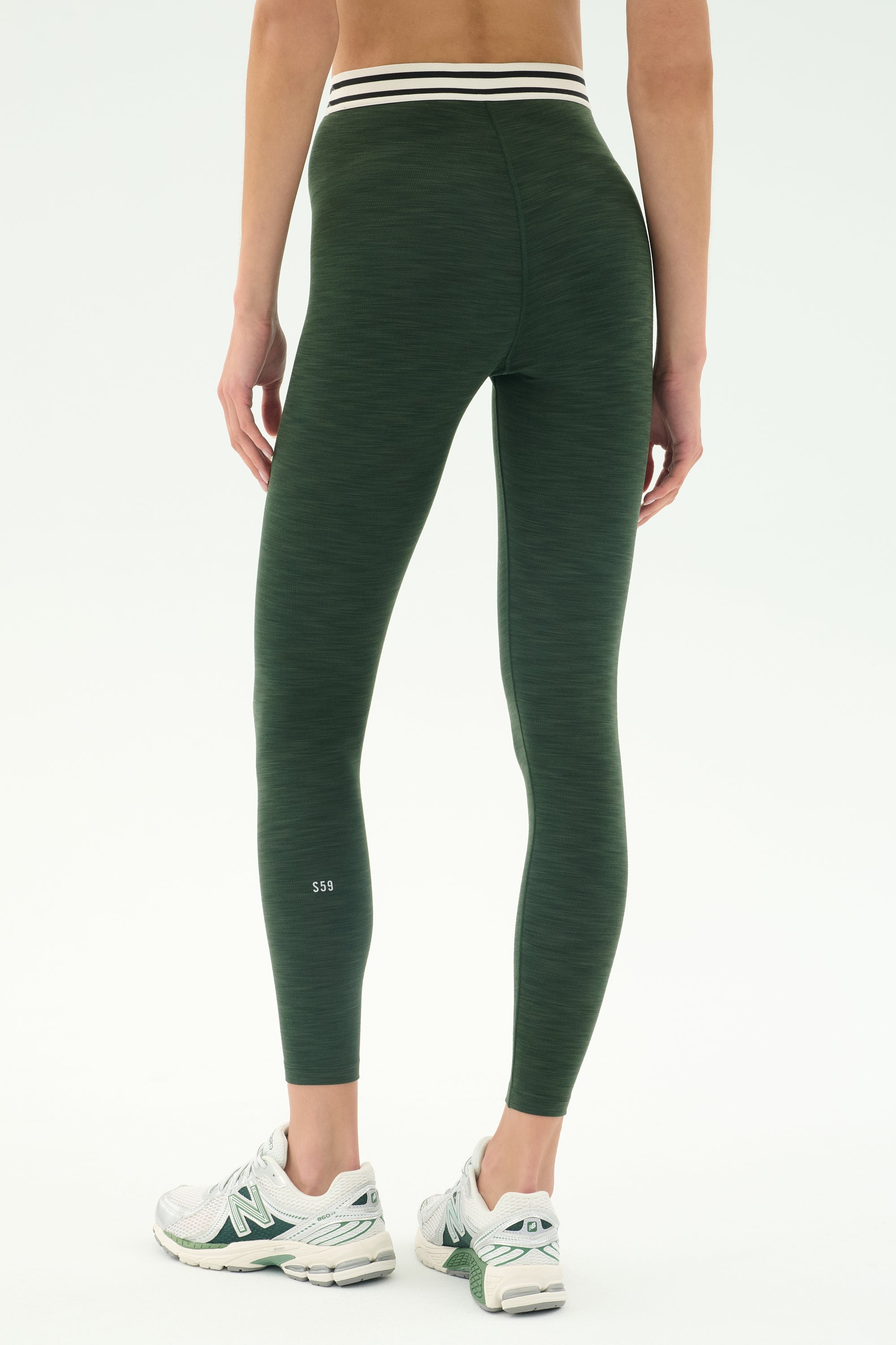 A person stands on a white background wearing SPLITS59 Bailey Active Rib High Waist 7/8 leggings in Heather Military with a striped waistband and white athletic sneakers—ideal for gym workouts.