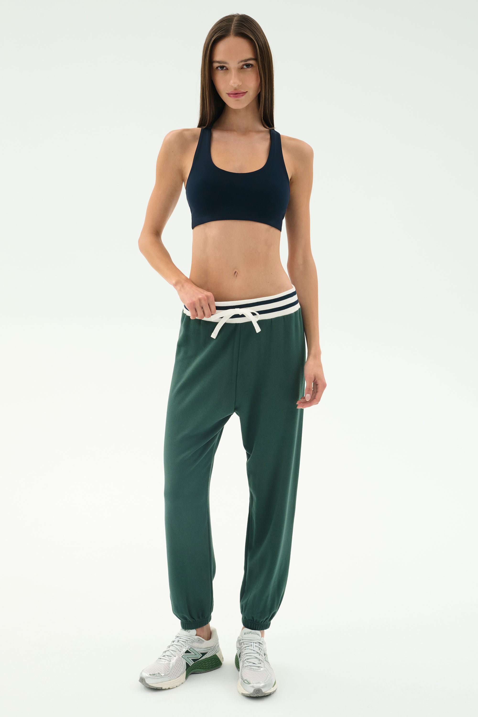 Woman standing against a plain background, wearing a black spandex sports bra, SPLITS59 Andie Oversized Fleece Sweatpant W/ Stripe in Military, and white sneakers.