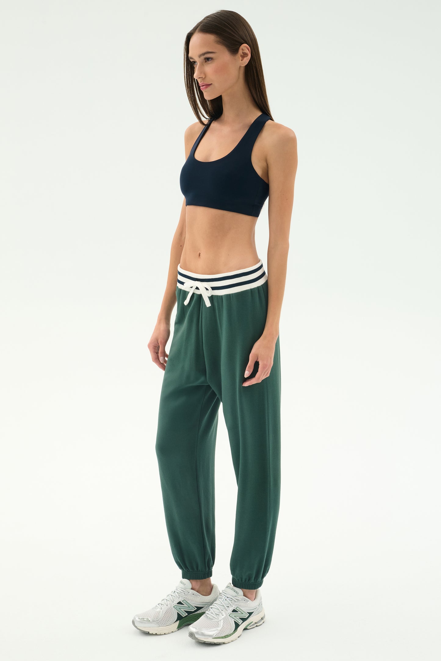 A woman in profile wears the SPLITS59 Andie Oversized Fleece Sweatpant W/ Stripe - Military, paired with a navy spandex sports bra and white sneakers, set against a plain white background.
