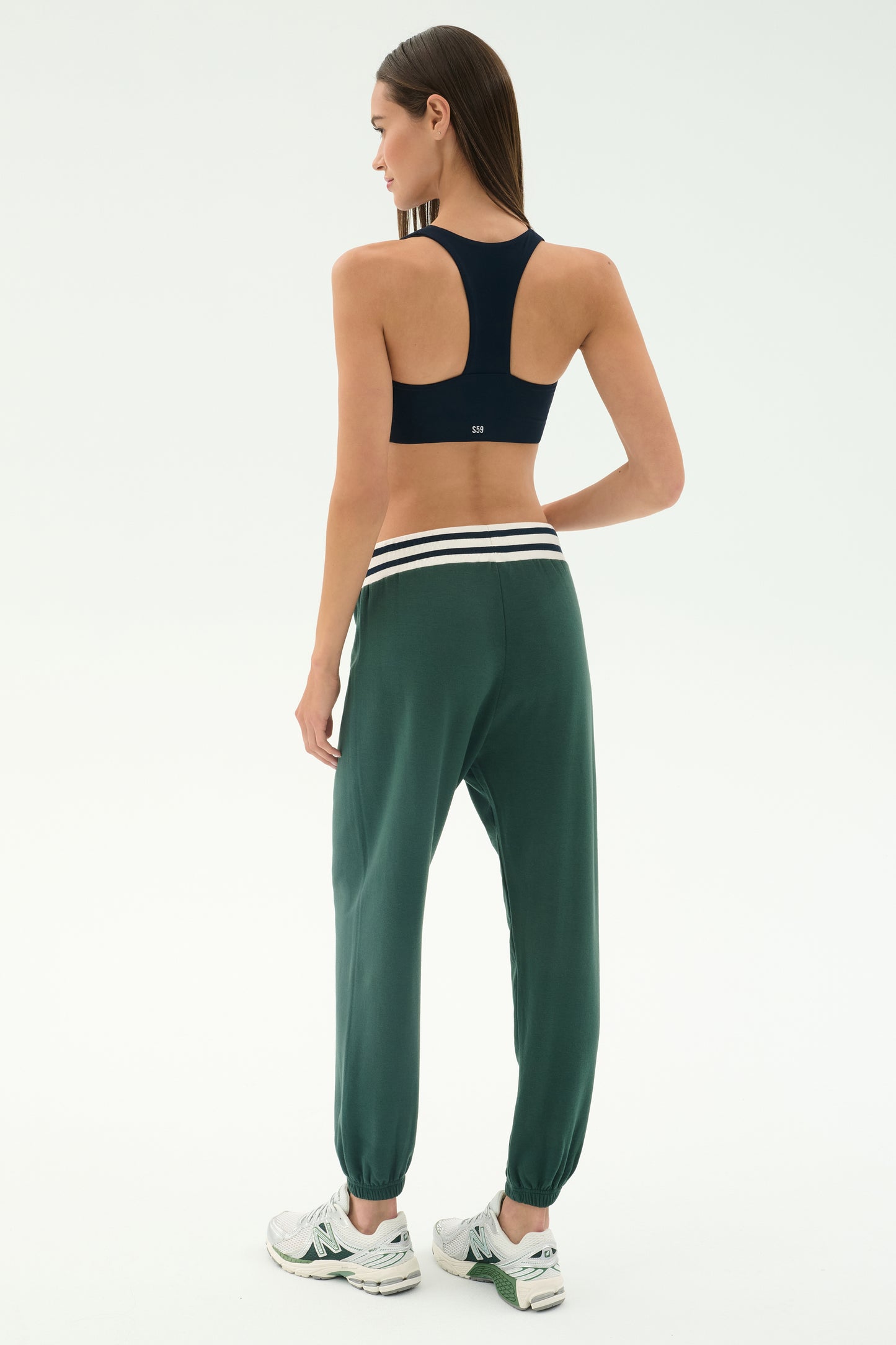 A woman faces away, wearing a black sports bra, SPLITS59 Andie Oversized Fleece Sweatpant w/ Stripe in Military, and white sneakers against a plain white background.