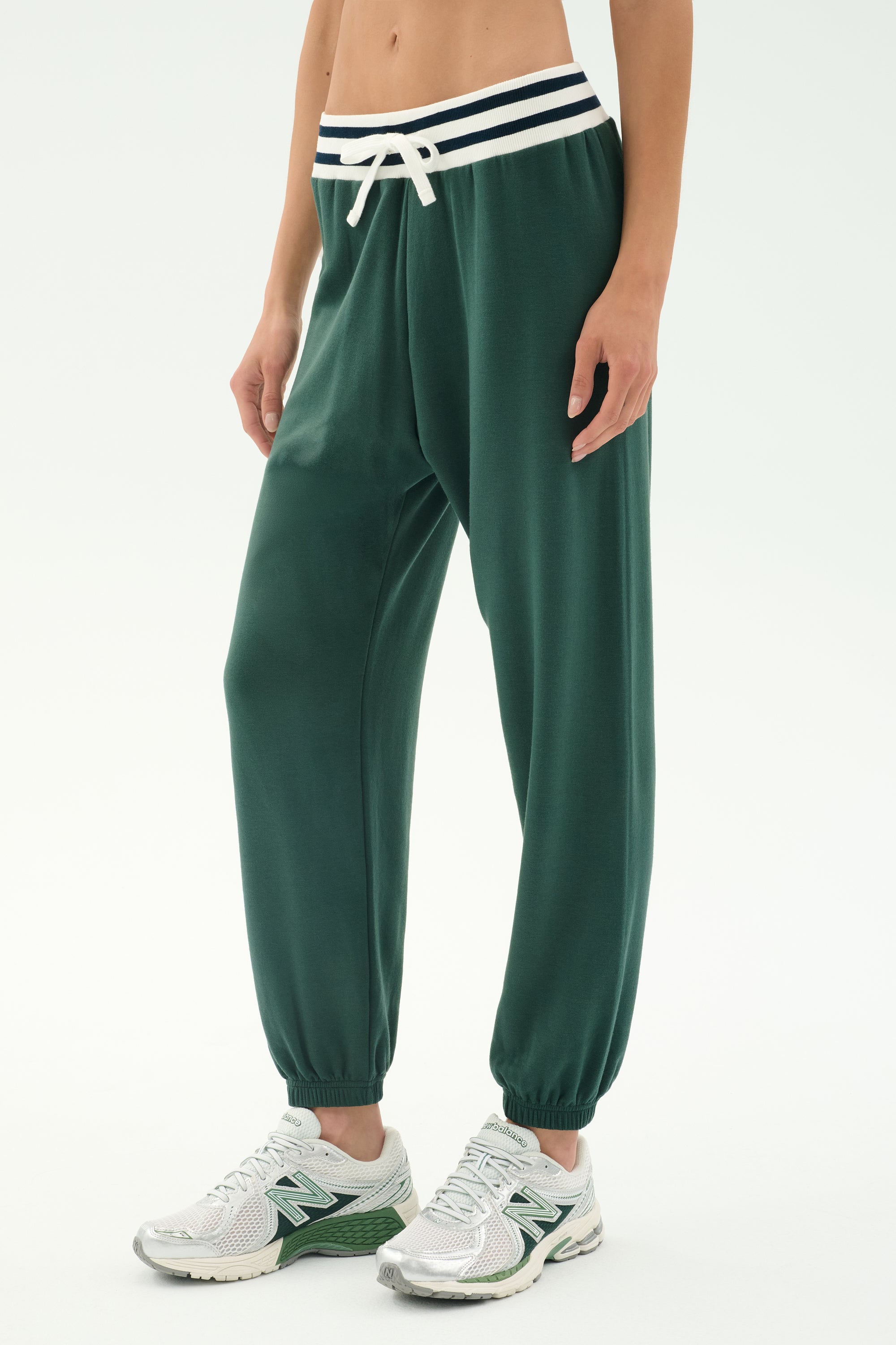 Model wears SPLITS59 Andie Oversized Fleece Sweatpant W/ Stripe - Military, featuring a soft spandex blend, white drawstring, and striped waistband, paired with white sneakers against a plain light background.