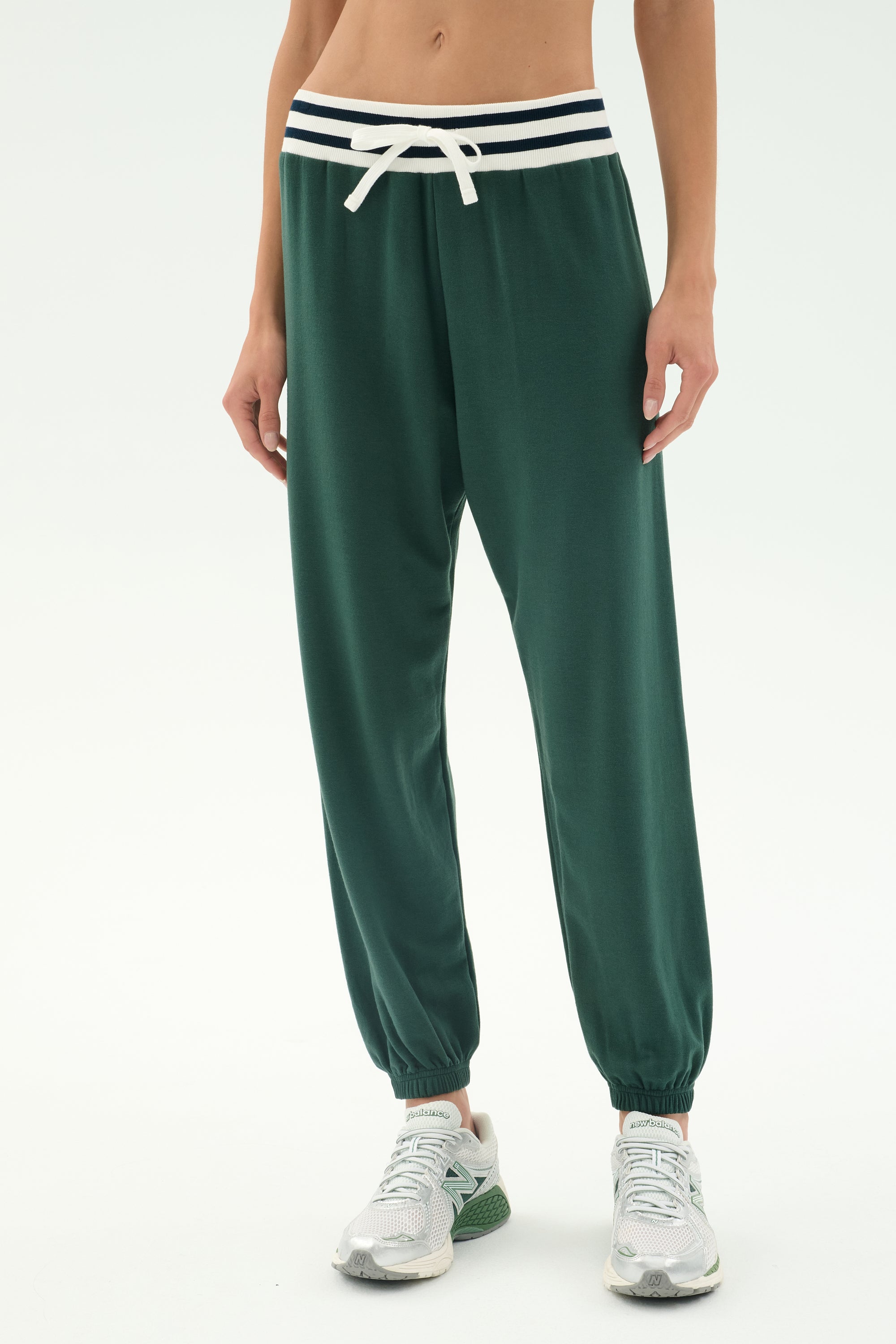 A person wearing SPLITS59 Andie Oversized Fleece Sweatpant W/ Stripe in Military, featuring a striped waistband and white drawstring, paired with white and green athletic shoes, stands against a plain background.