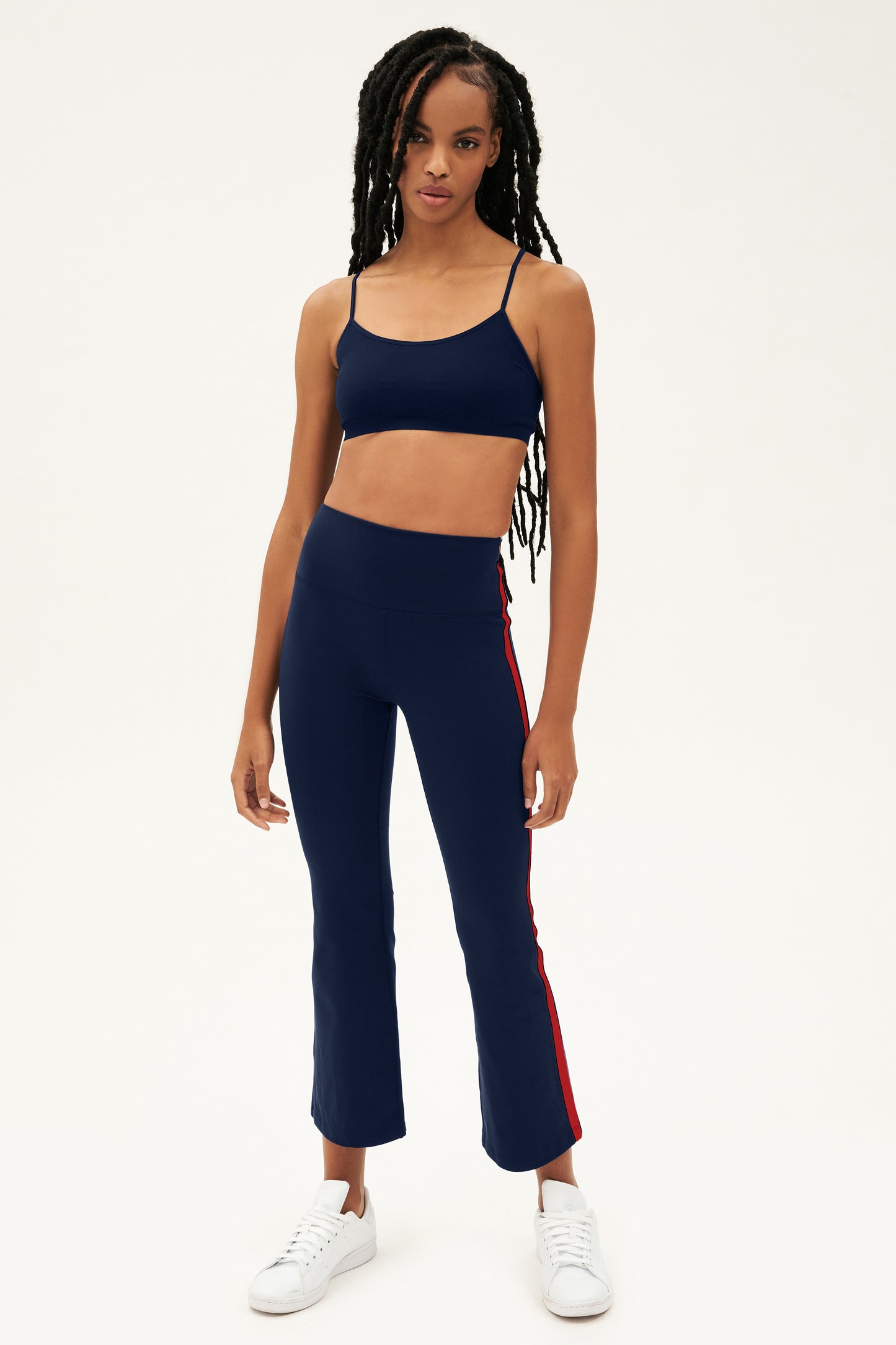 Raquel wears the SPLITS59 Raquel High Waist Supplex Crop in Indigo/Pirate Red, featuring 4-way stretch supplex for yoga, paired with white sneakers and standing against a plain white background.