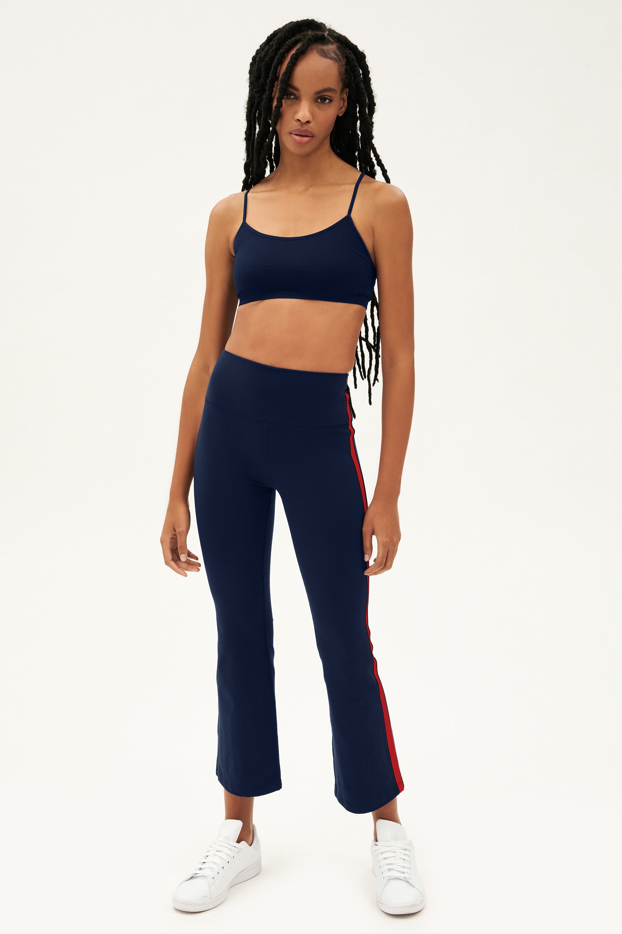 Raquel wears the SPLITS59 Raquel High Waist Supplex Crop in Indigo/Pirate Red, featuring 4-way stretch supplex for yoga, paired with white sneakers and standing against a plain white background.