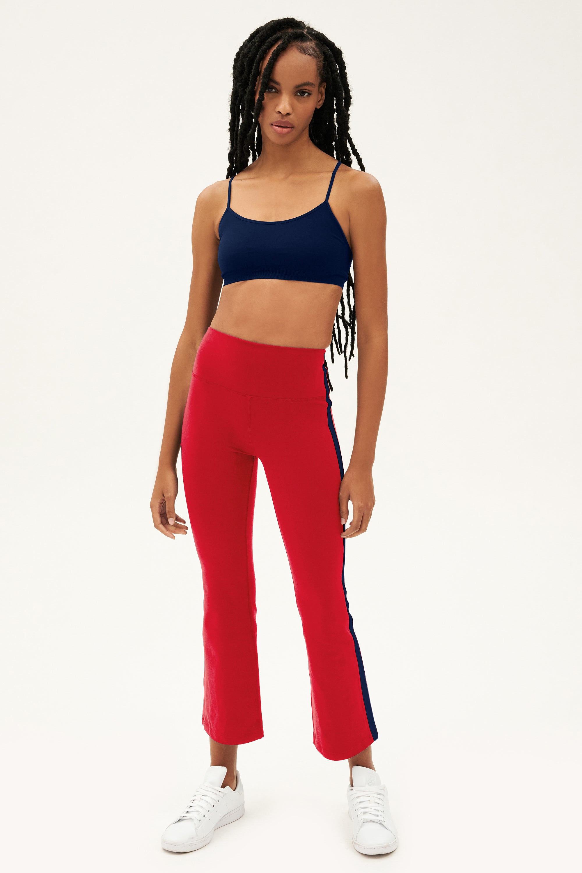 A woman stands against a plain background wearing SPLITS59’s Raquel High Waist Crop pants in Pirate Red/Indigo, featuring navy side stripes and 4-way stretch fabric, styled with a navy crop top and white sneakers.