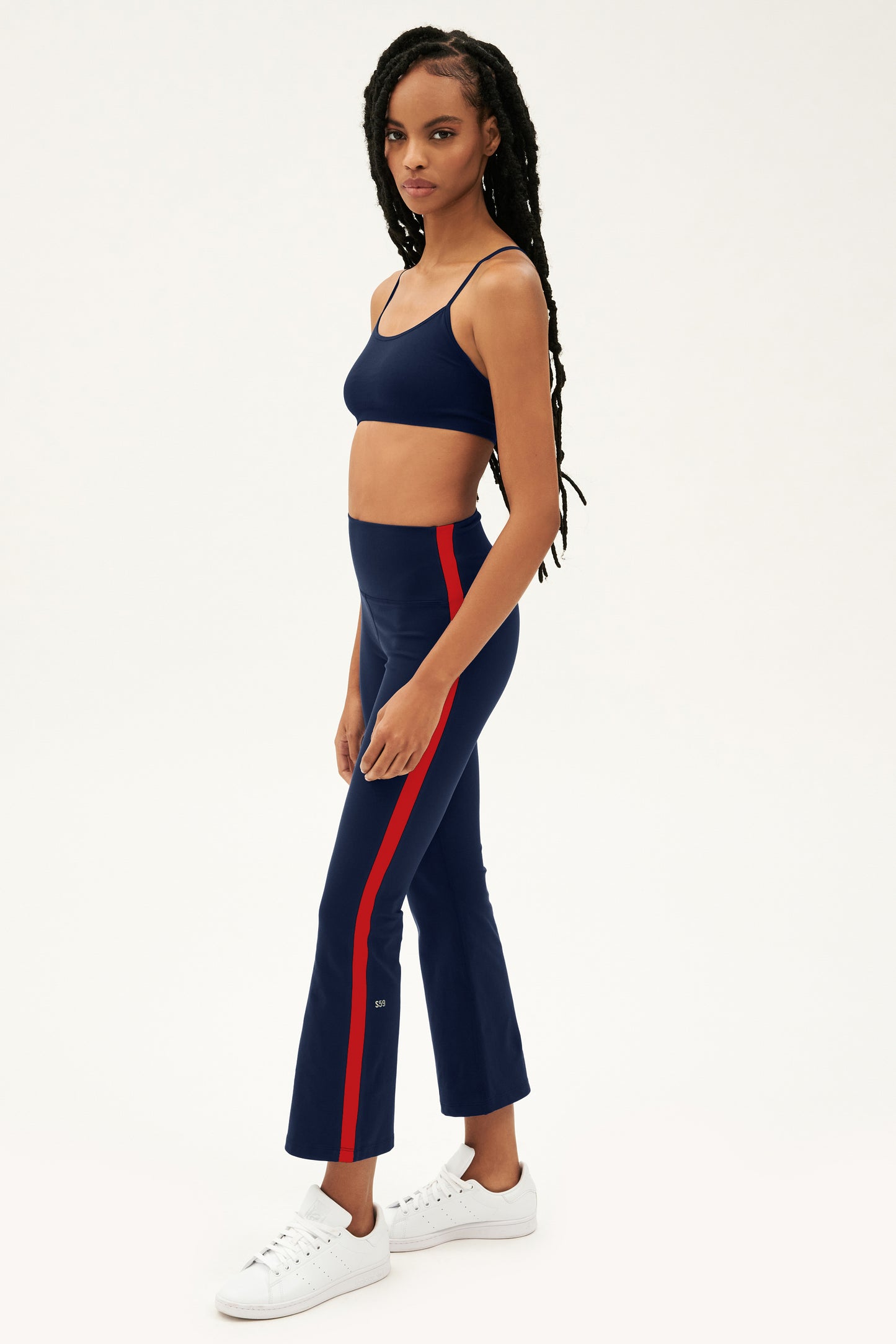 Raquel models the SPLITS59 Raquel High Waist Supplex Crop in Indigo/Pirate Red—navy high-waist crops with red side stripes, made from 4-way stretch supplex, perfect for yoga—styled with white sneakers against a plain background.