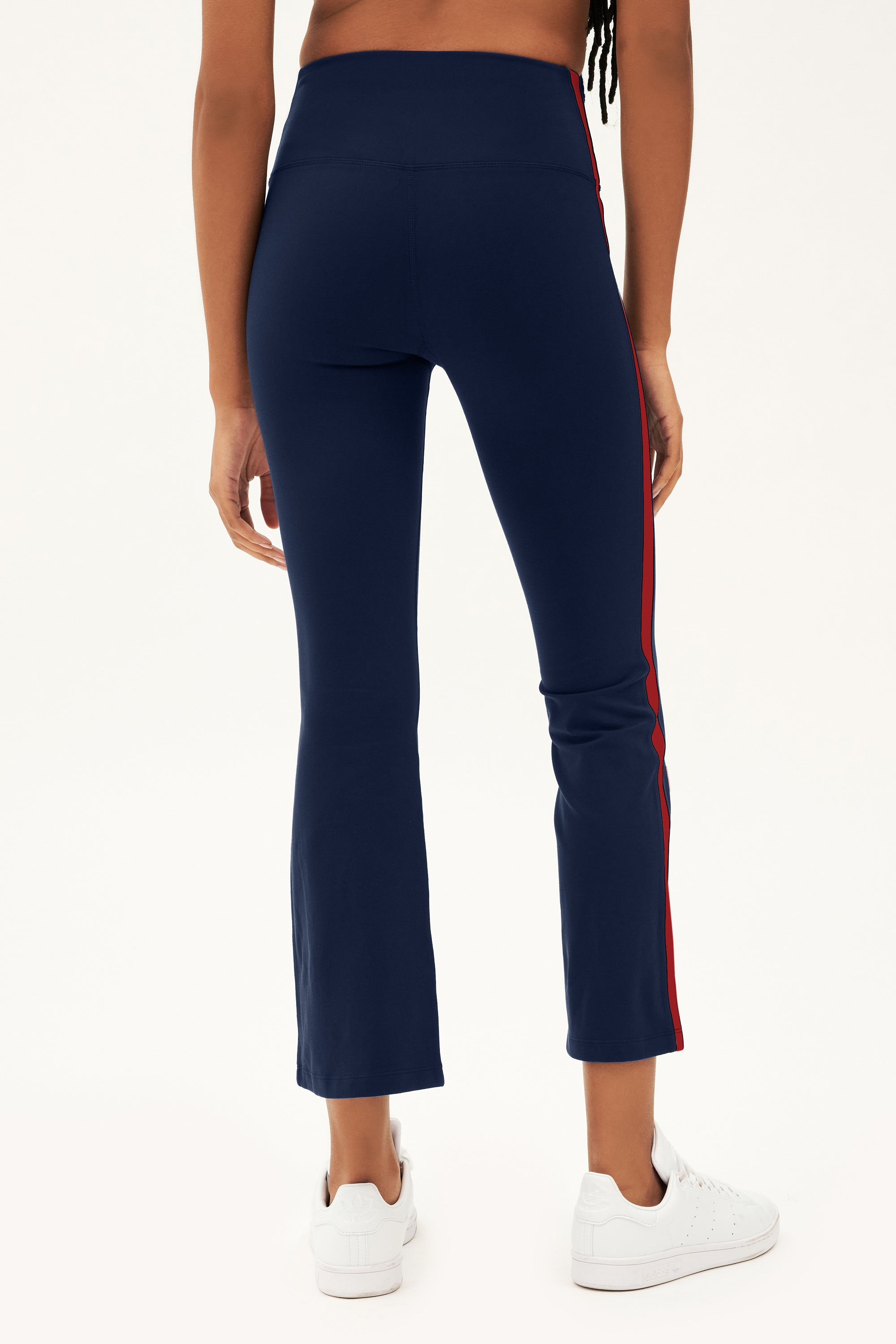 A woman faces away, wearing SPLITS59 Raquel High Waist Crop pants in Indigo/Pirate Red with white sneakers, set against a plain background.
