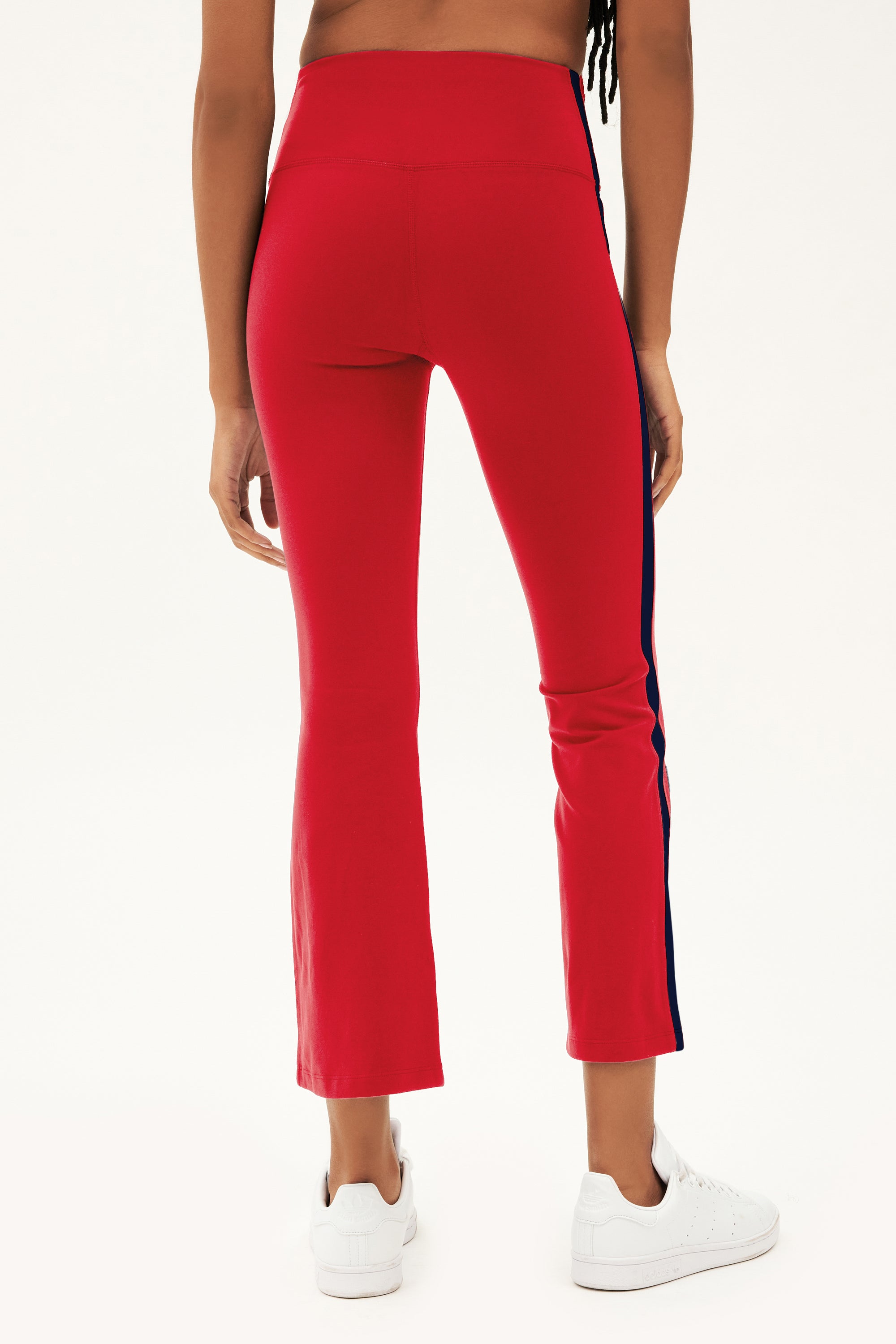 A person wears SPLITS59 Raquel High Waist Crop pants in Pirate Red/Indigo, featuring a side stripe and 4-way stretch, with white sneakers, shown from the back against a plain background.