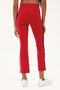 A person wears SPLITS59 Raquel High Waist Crop pants in Pirate Red/Indigo, featuring a side stripe and 4-way stretch, with white sneakers, shown from the back against a plain background.