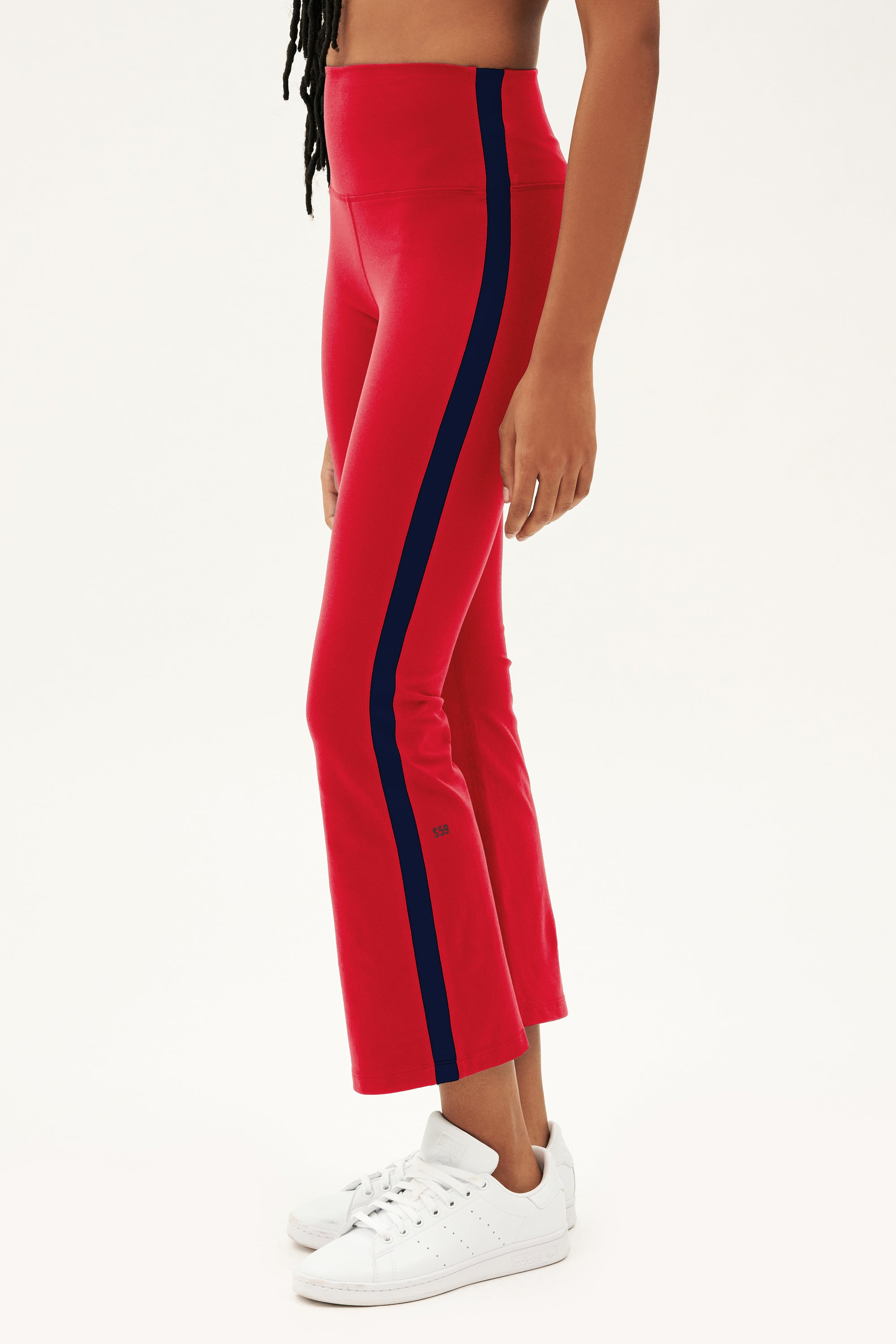 A person wearing SPLITS59 Raquel High Waist Supplex Crop leggings in Pirate Red/Indigo with white sneakers stands against a plain white background.