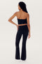 A person with long braids is standing with their back to the camera, wearing a strapless black top (Hazel Airweight Crop Bandeau - Black by SPLITS59), black flared pants made of spandex, and white sneakers against a plain white background.