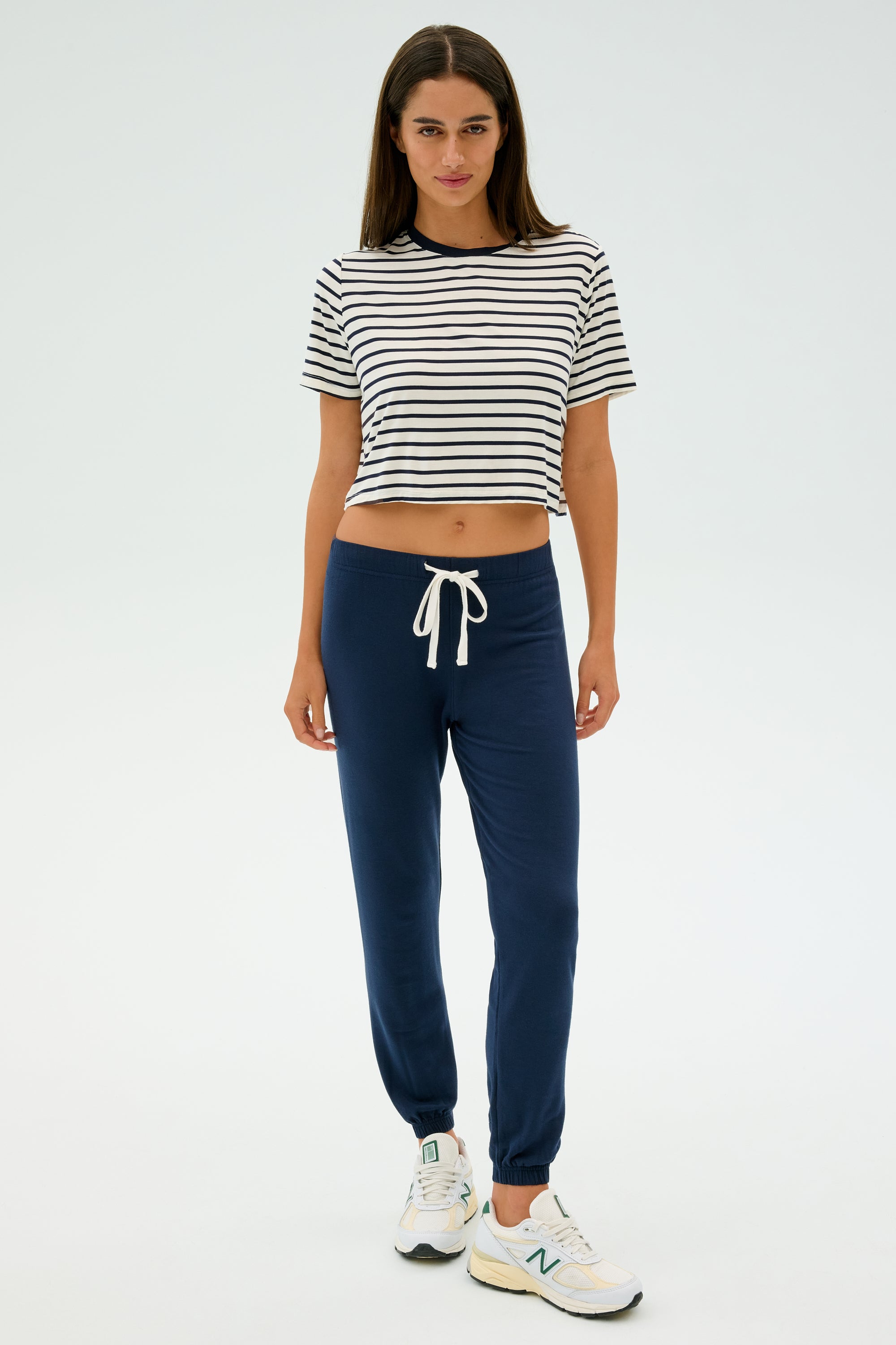 A woman stands on a plain backdrop in a striped crop top, navy joggers, and white sneakers, ready for cool weather workouts. She embraces SPLITS59's Sonja Fleece Sweatpant in Indigo, effortlessly combining style with comfort.