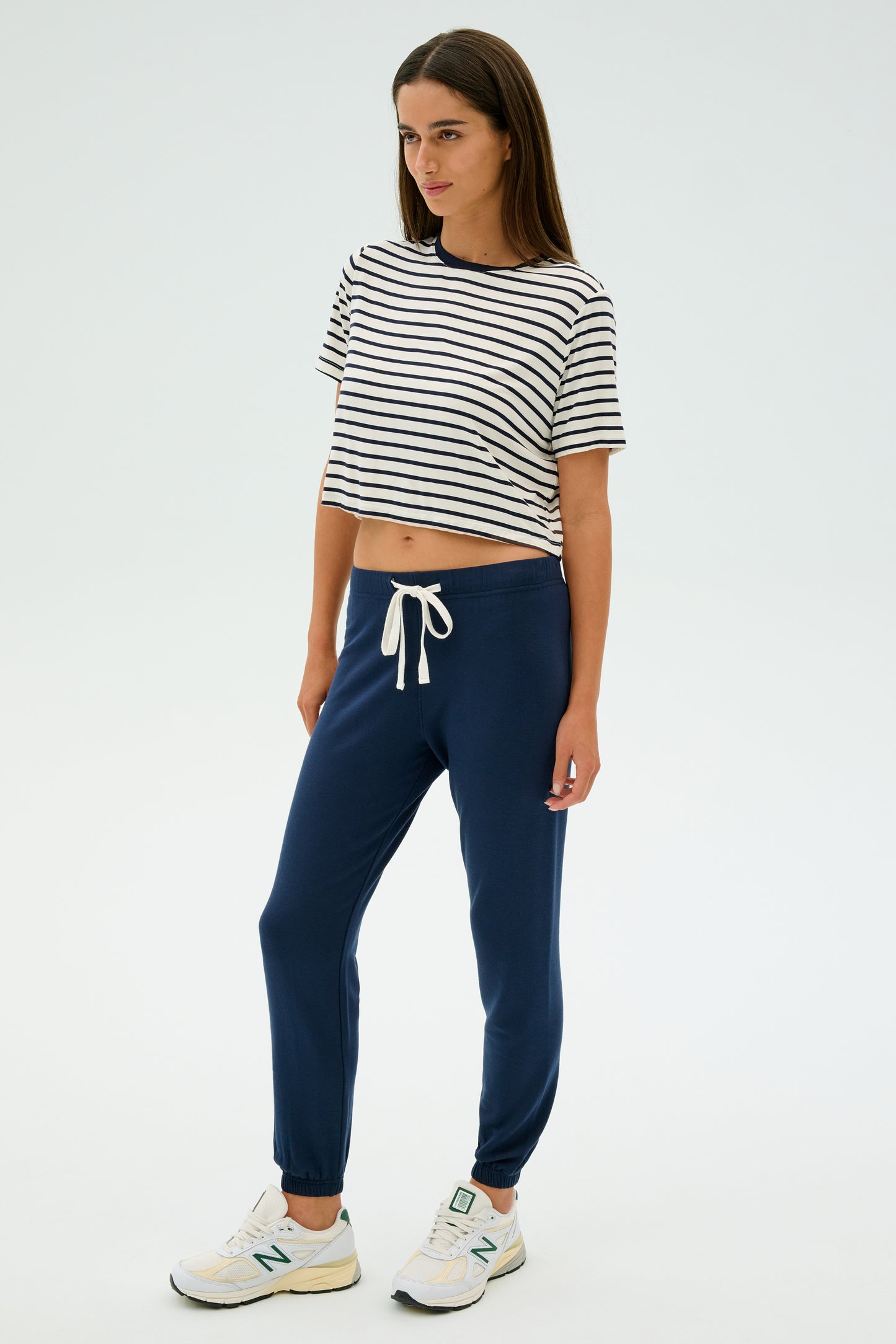 A person models SPLITS59's Sonja Fleece Sweatpant in Indigo, paired with a striped T-shirt and white sneakers, posed against a plain background. They look to the side, perfectly styled for cool-weather workouts.