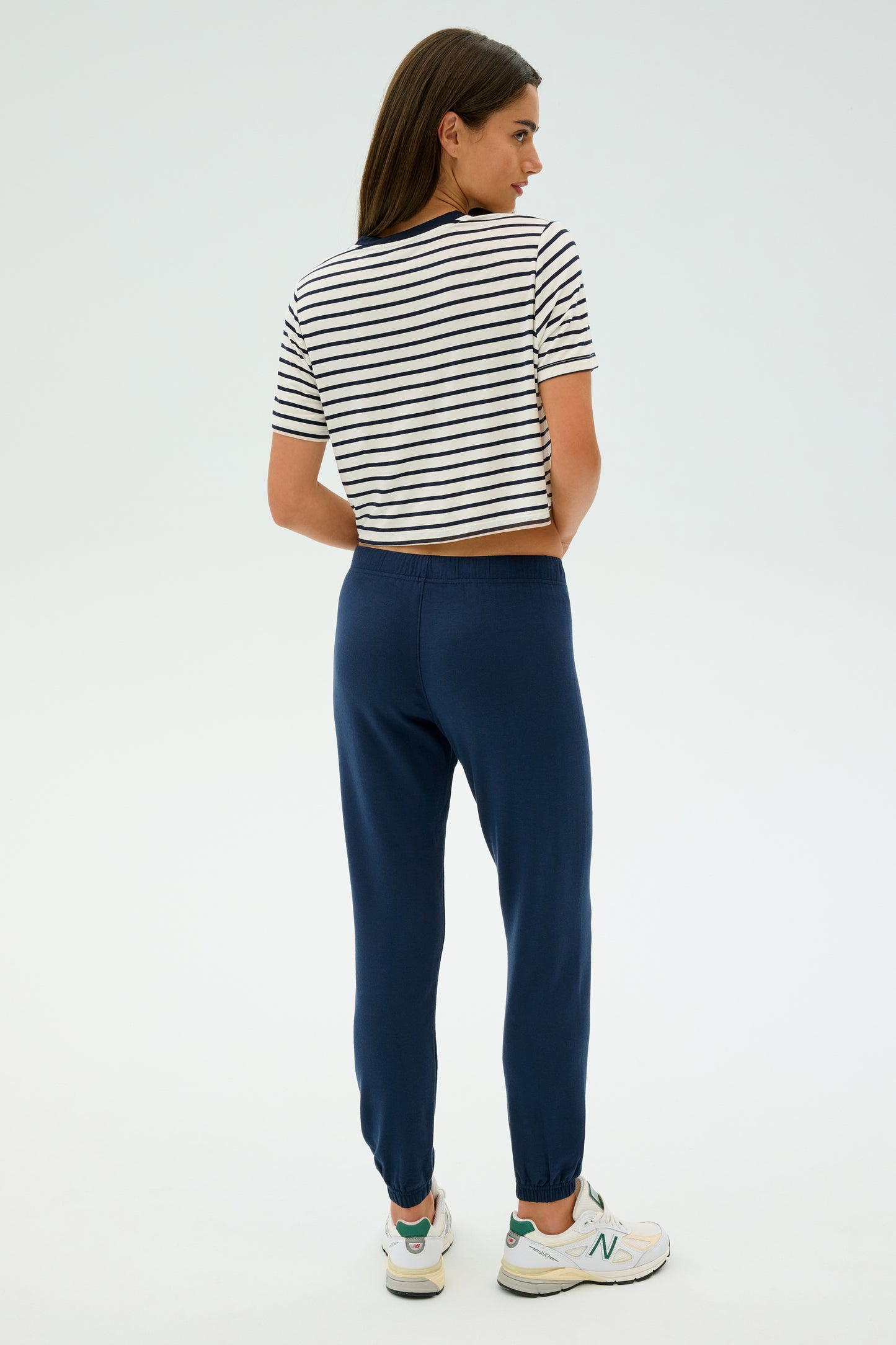 A woman with long hair stands facing away in a striped T-shirt, SPLITS59's Sonja Fleece Sweatpant in Indigo, and white sneakers against a plain background.