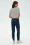 A woman with long hair stands facing away in a striped T-shirt, SPLITS59's Sonja Fleece Sweatpant in Indigo, and white sneakers against a plain background.